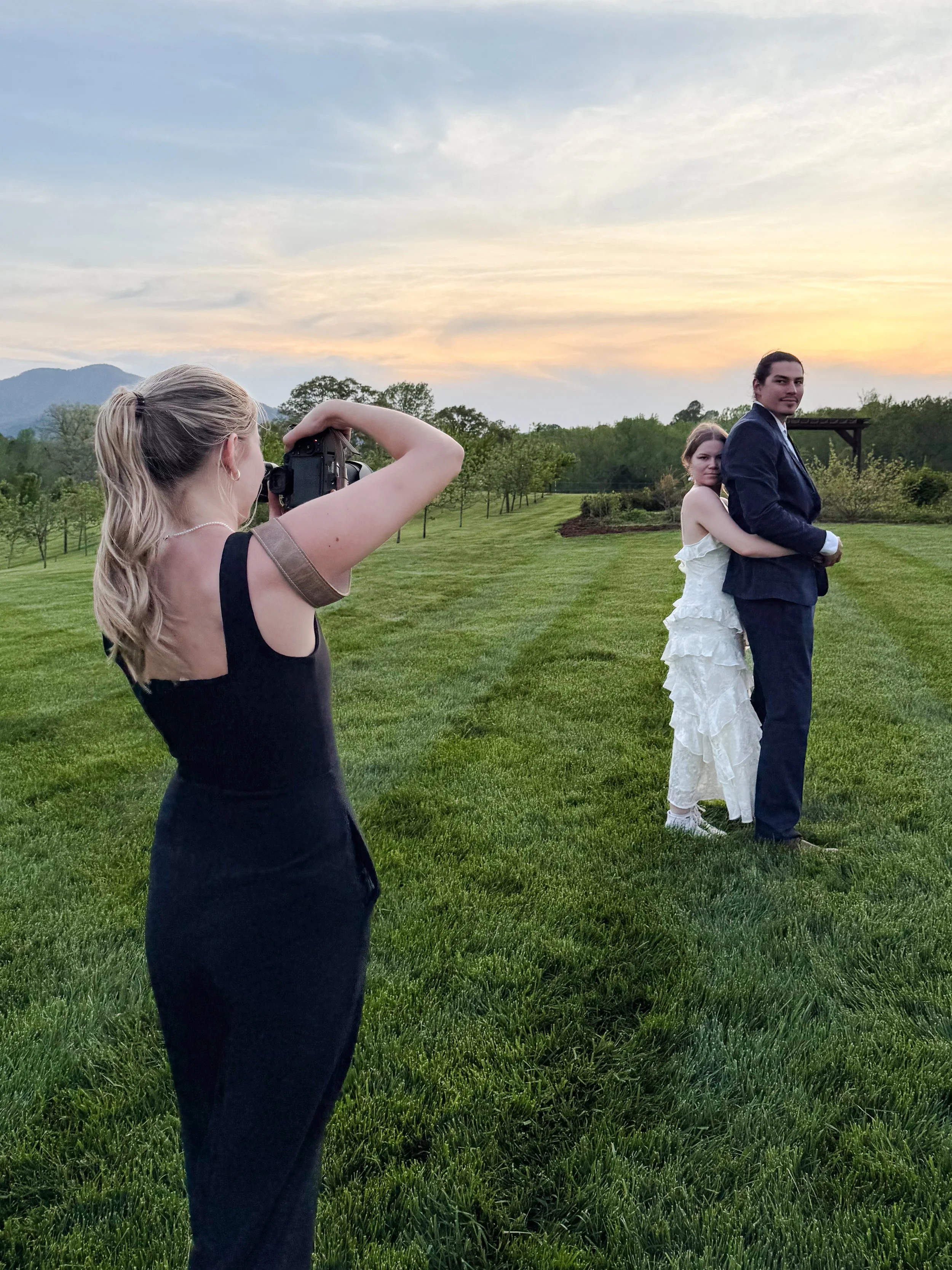 A photographer takes a picture of a couple embracing outdoors during sunset, with green grass, trees, and mountains in the background.