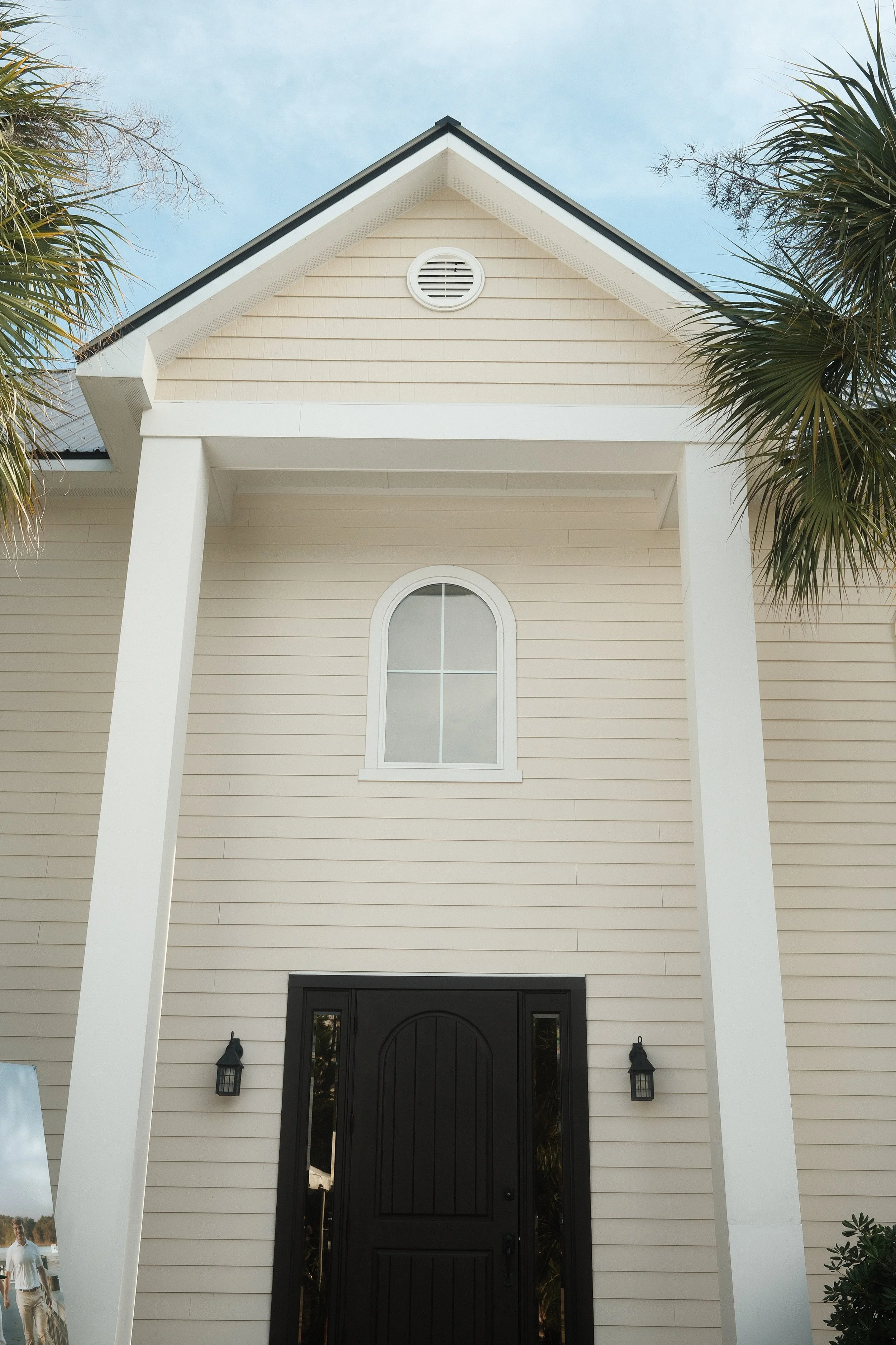 Front view of a house with black door, beige siding, columns, and palm trees on sides, against a blue sky.