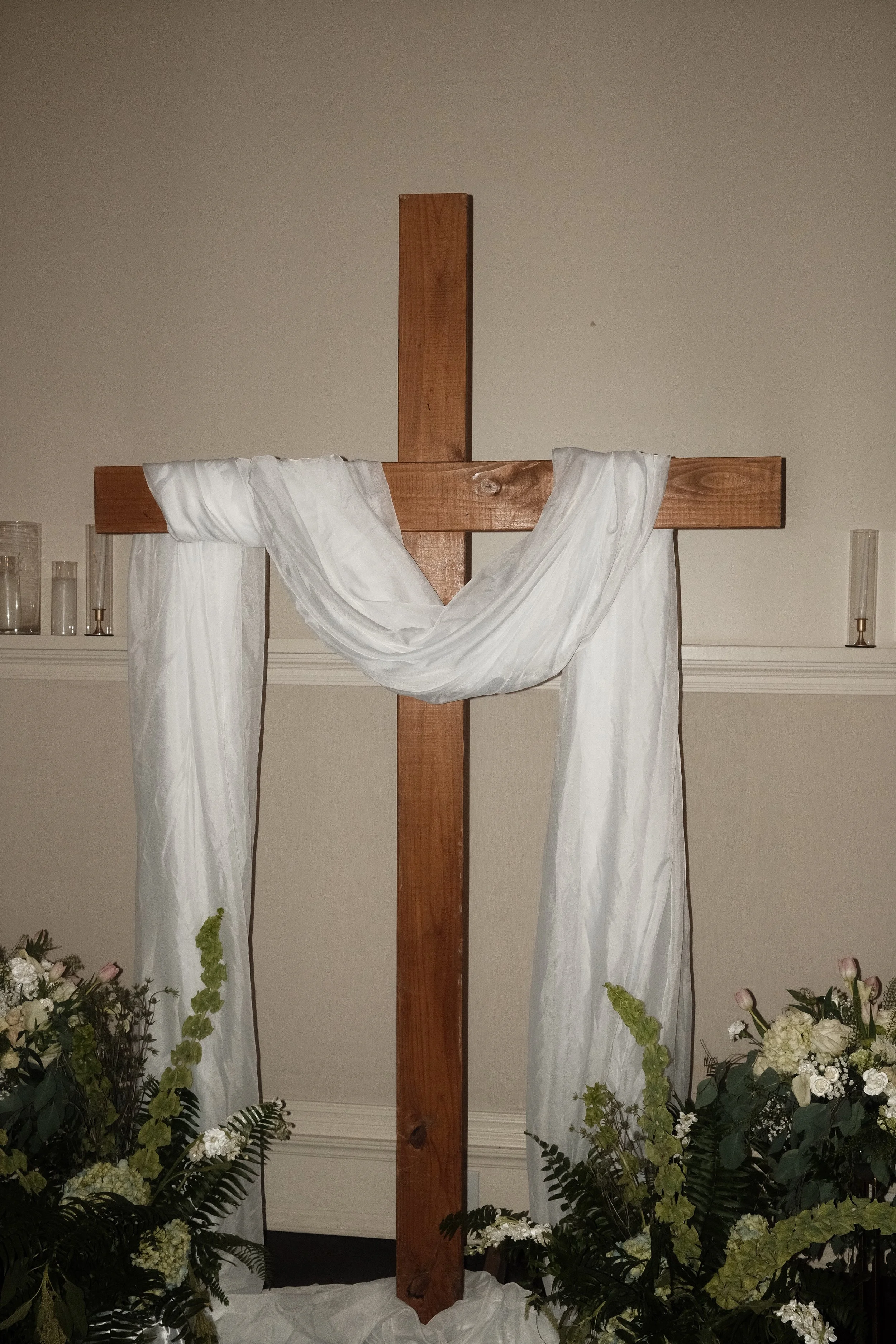 A wooden cross draped with white cloth, surrounded by floral arrangements, set against a plain wall.