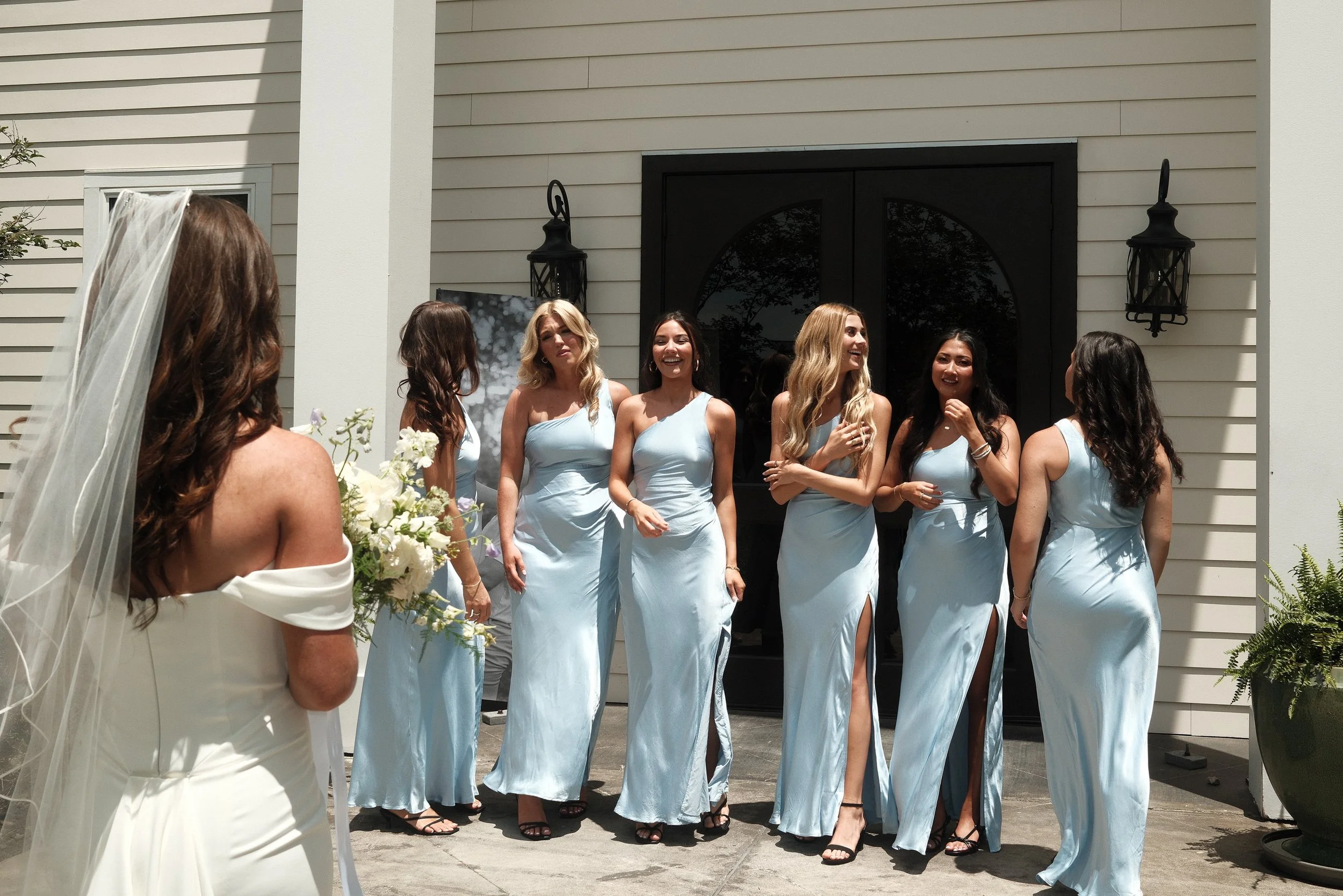 Group of women in matching light blue dresses standing outside a house, smiling and chatting during a daytime event.