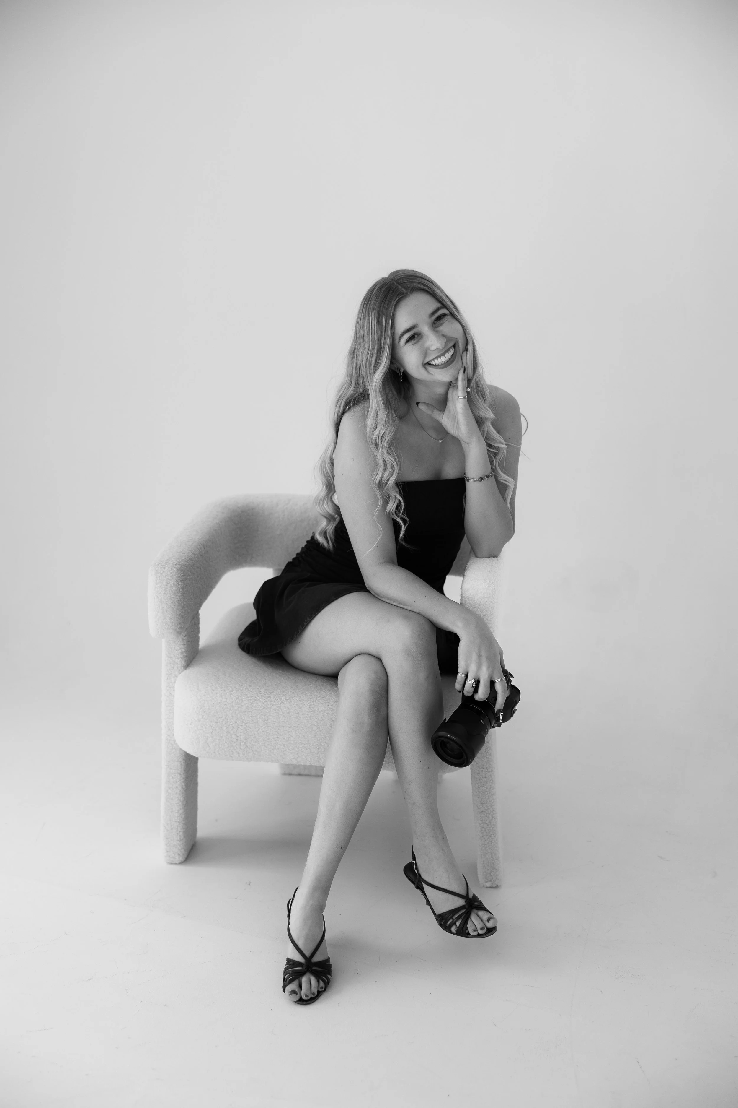 A woman with long wavy hair, wearing a strapless top and a skirt, sitting on a chair, and smiling while holding a camera.
