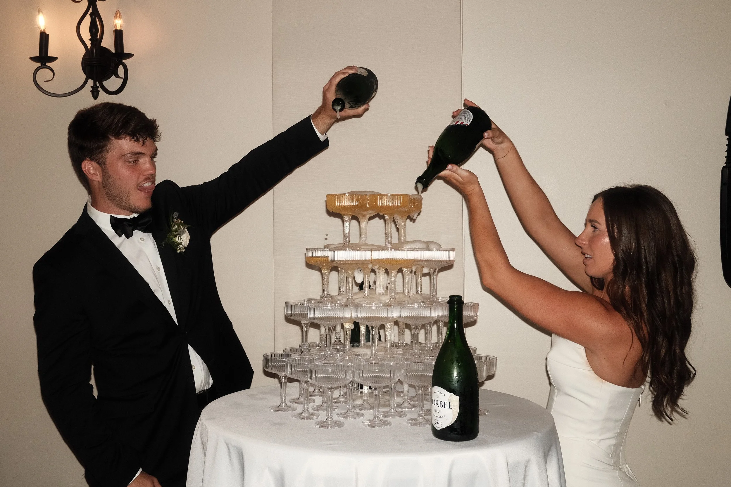 A man in a tuxedo and a woman in a white dress are raising bottles of champagne over a champagne tower at a celebration event.