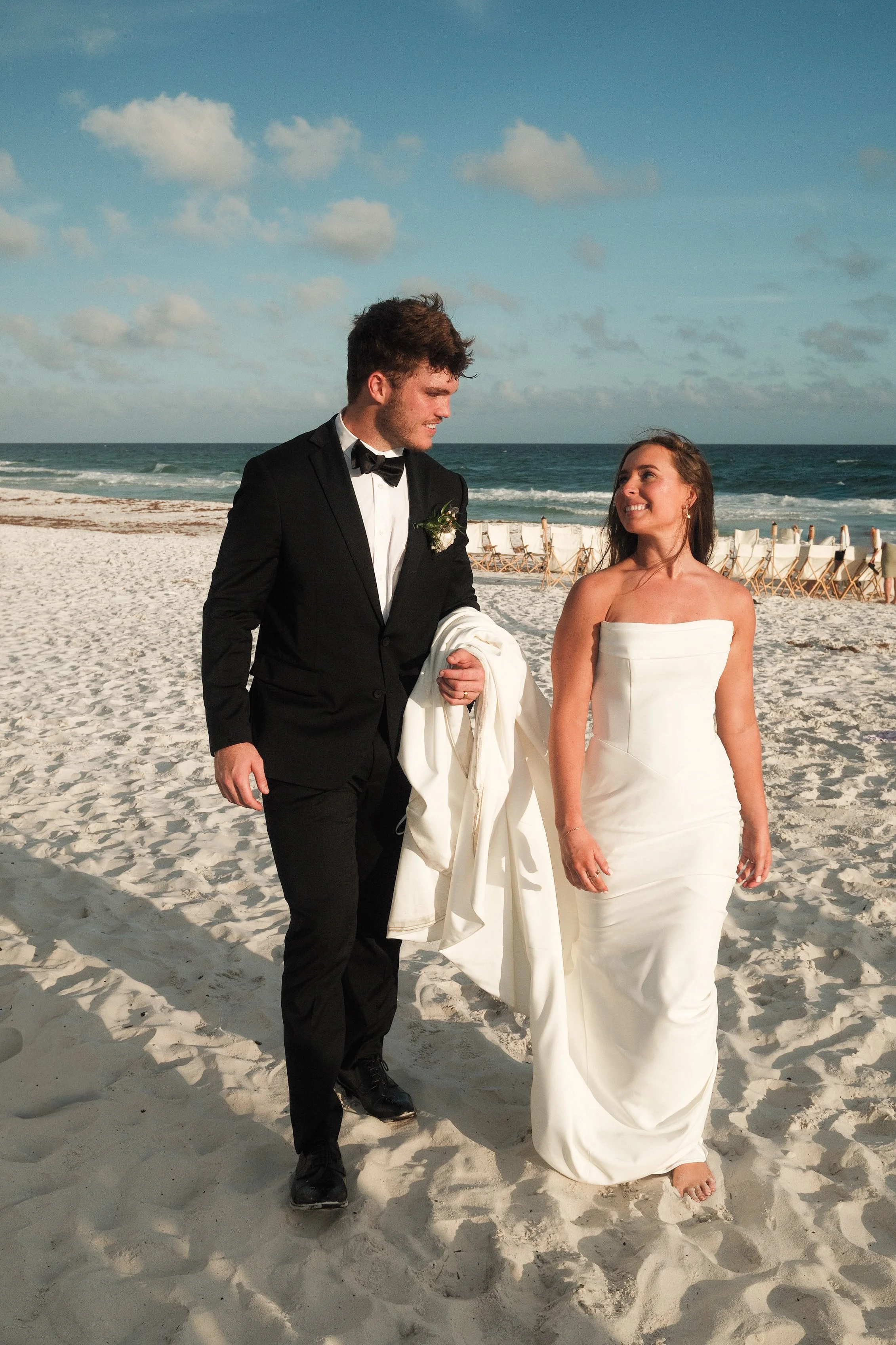 A man in a black tuxedo and a woman in a white strapless wedding gown walking on a sandy beach, holding hands and smiling at each other with the ocean and a line of chairs in the background.