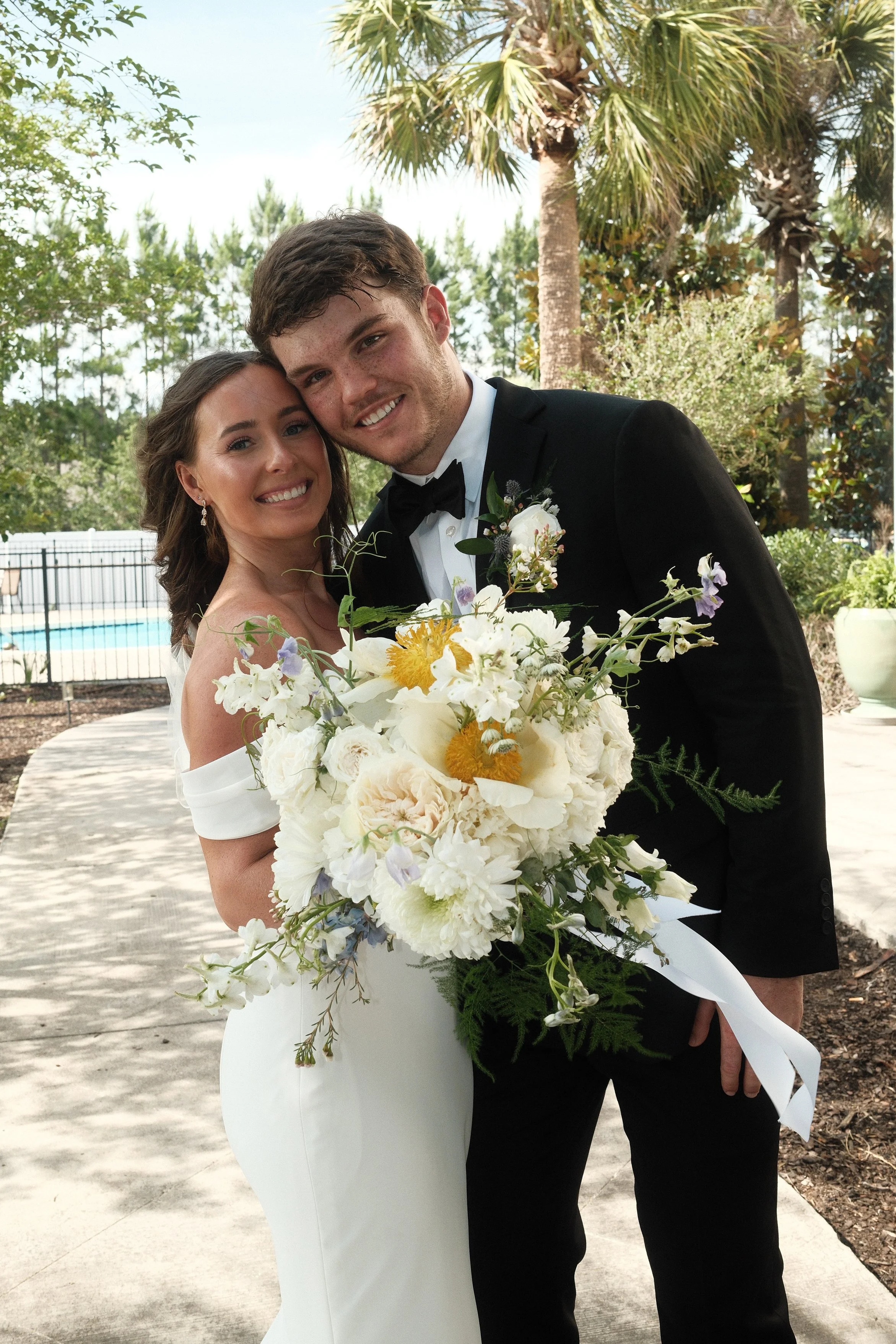 A bride and groom smiling and hugging each other on their wedding day outdoors, with trees and a pool in the background. The bride is holding a large bouquet of white, purple, and yellow flowers.