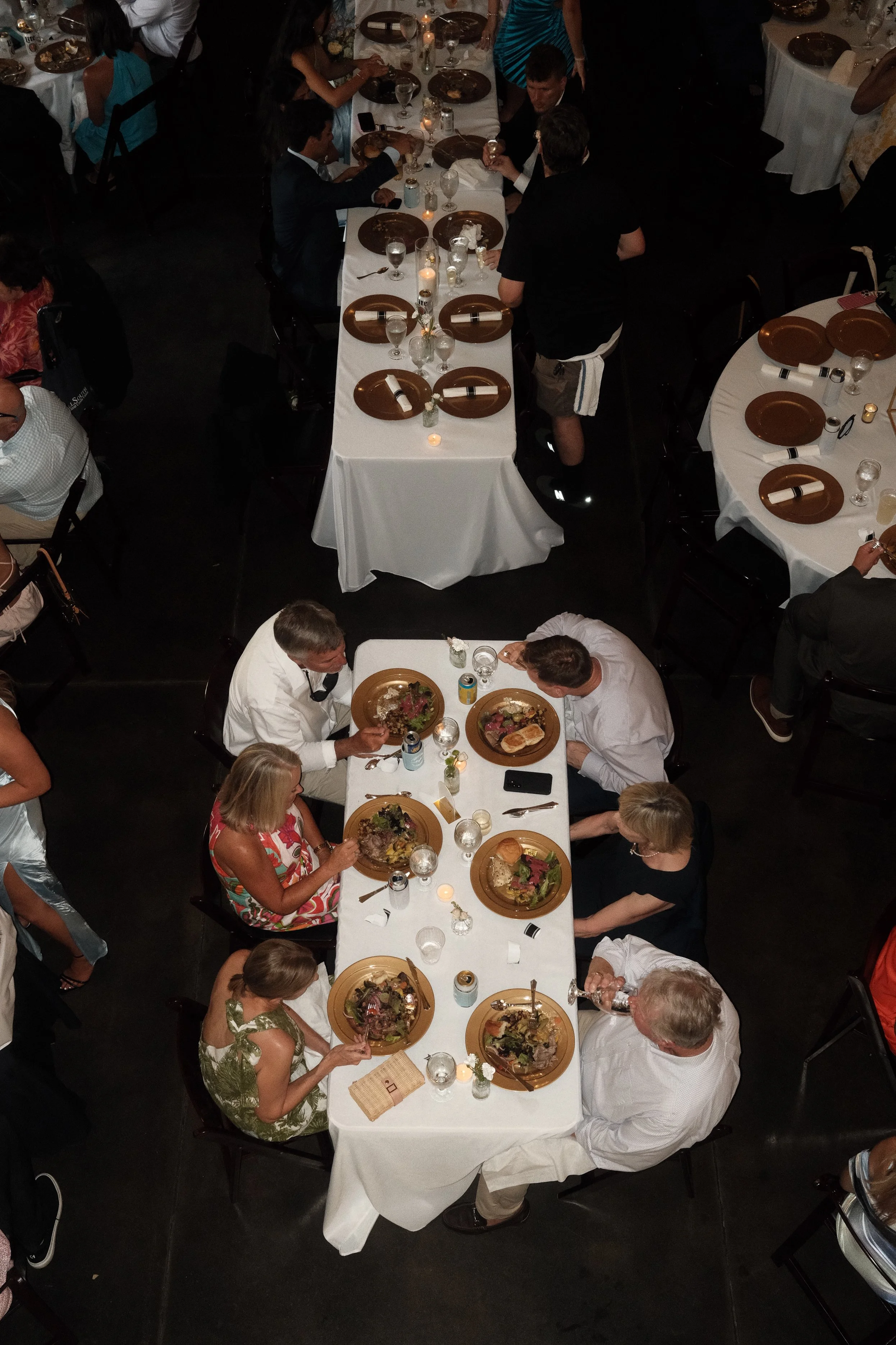 A large dining room filled with people at a formal event, seated around long tables with white tablecloths, gold plates, and glassware. The tables are decorated with small candles and floral arrangements, and many guests are eating or conversing.