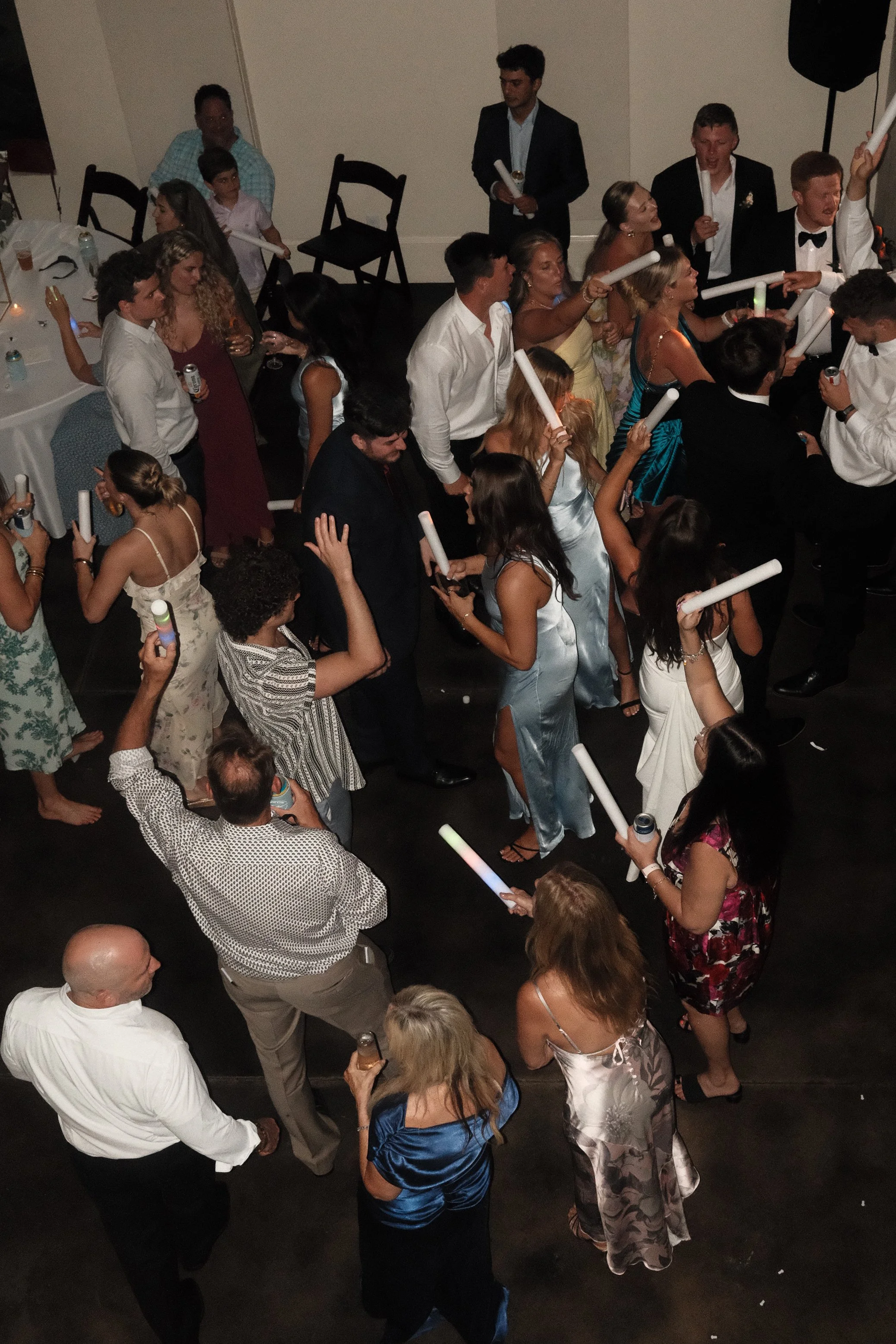 People dancing and celebrating at a wedding reception, holding glow sticks, with drinks on the tables.
