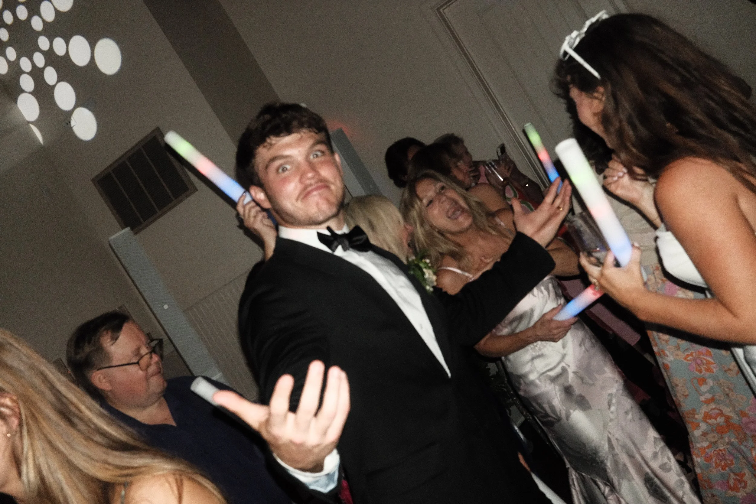 A man in a tuxedo holding a glow stick at a party with multiple women in elegant dresses, some holding glow sticks, in a room with a patterned carpet and wall decor.