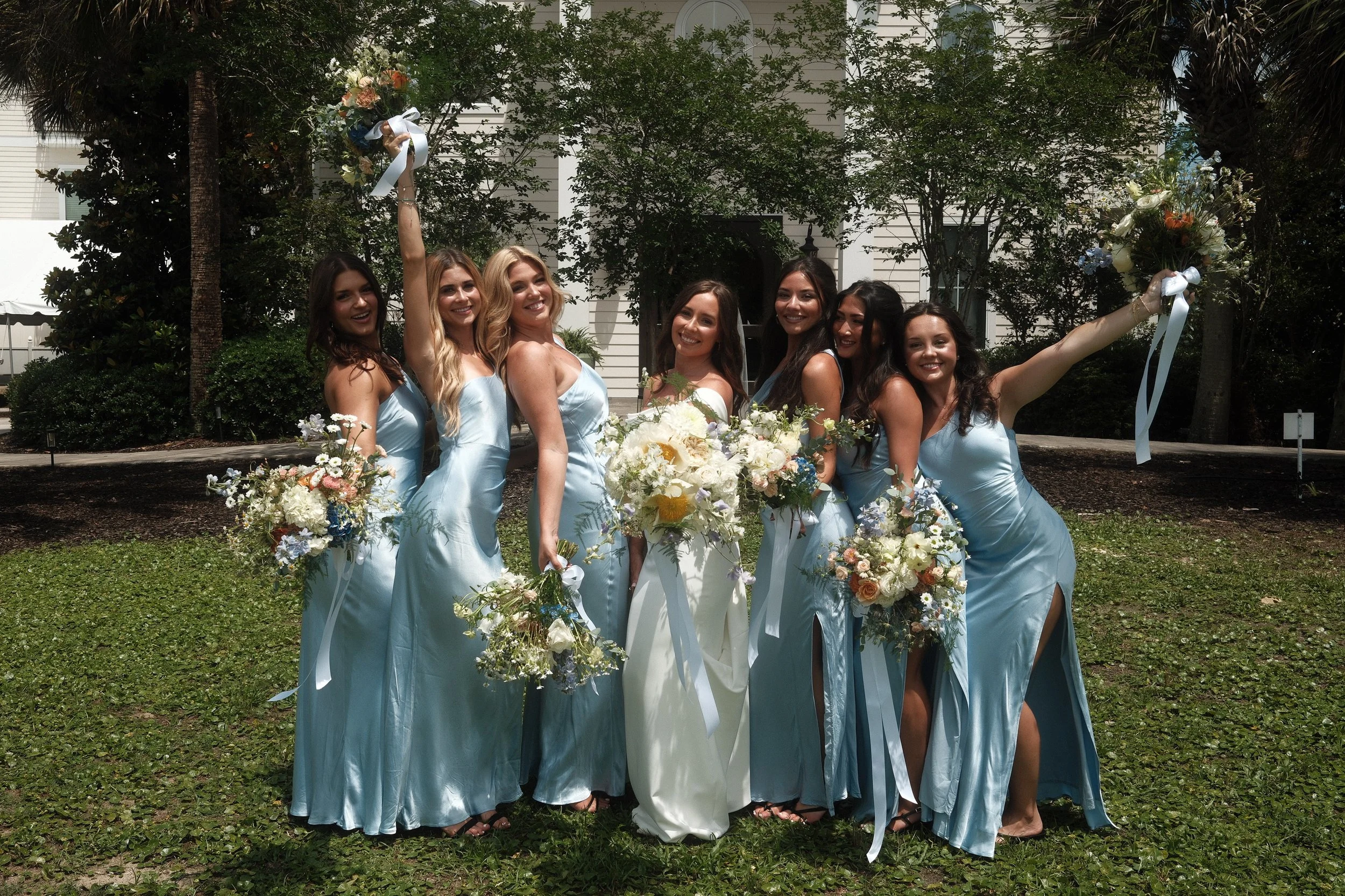 A group of six women dressed in matching light blue satin dresses, holding bouquets of flowers, celebrating outdoors on a sunny day. Two women are holding up bouquets, smiling and posing in front of a white house with trees and greenery.