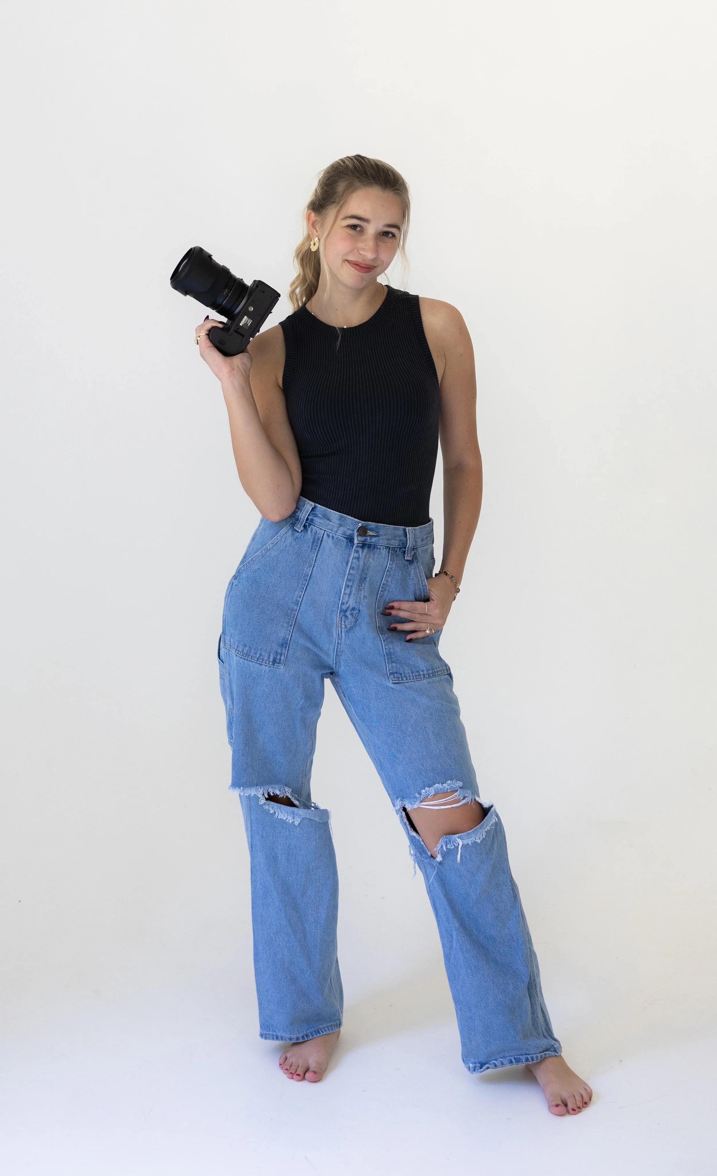 A woman with blonde hair in a ponytail, wearing a sleeveless black ribbed top and ripped blue jeans, standing barefoot against a white background, holding a professional camera in her right hand with a relaxed pose.