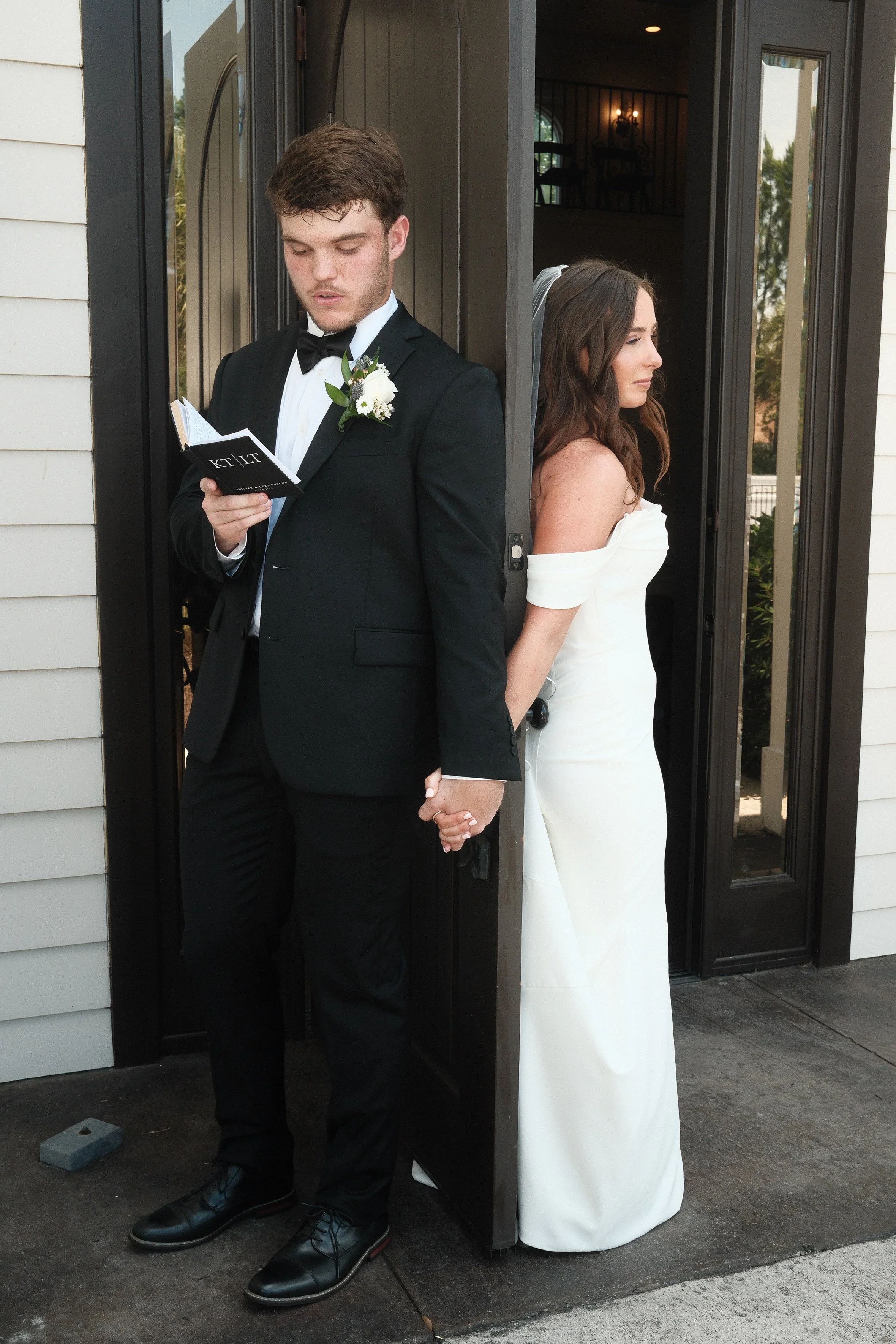 A newlywed couple standing back-to-back outside a doorway, holding hands. The groom is wearing a black tuxedo with a boutonniere, reading a small book. The bride is wearing a white off-shoulder gown, looking to the side.
