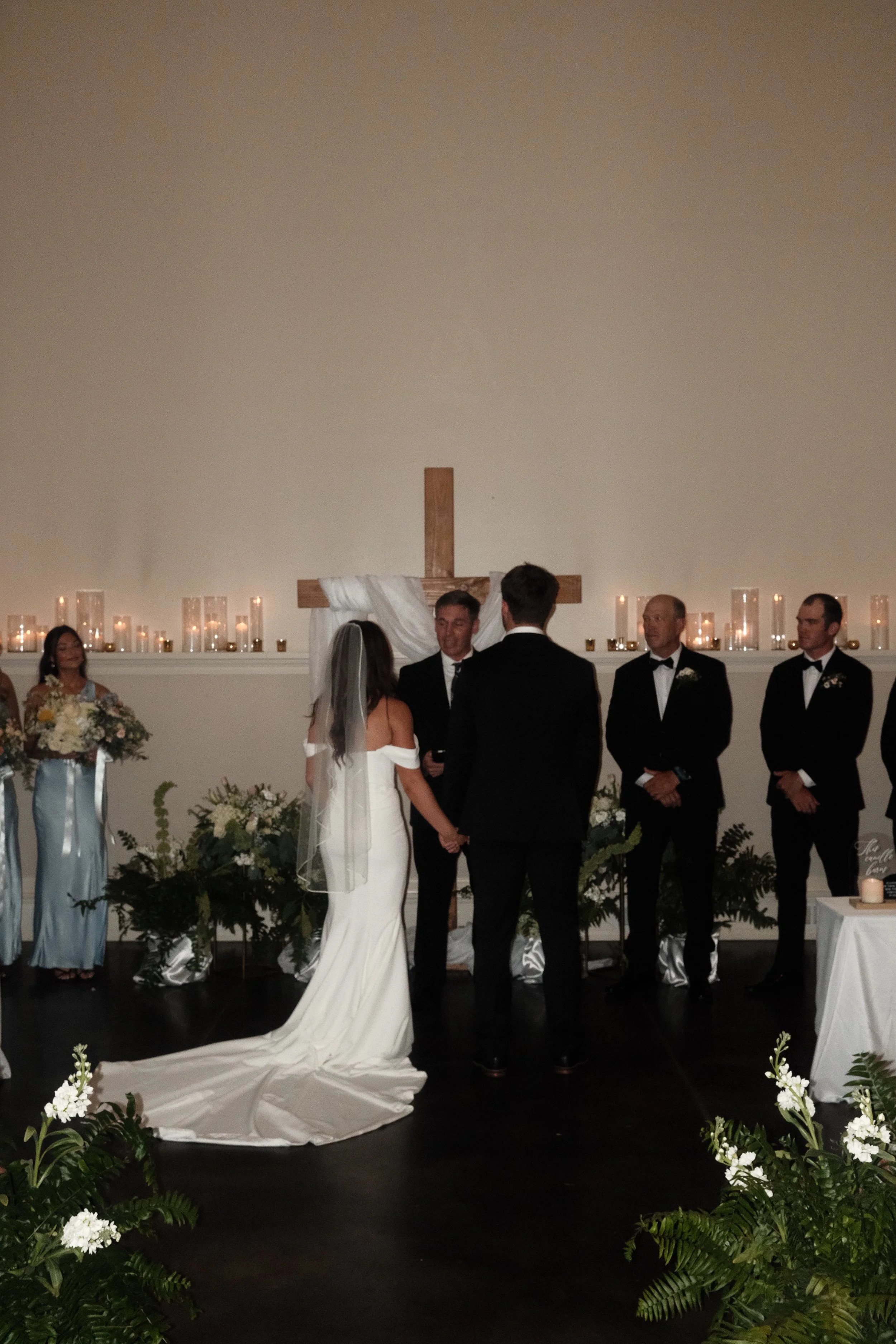 Wedding ceremony with bride and groom holding hands, officiant and guests in formal attire, decorated with candles, flowers, and a cross in the background.