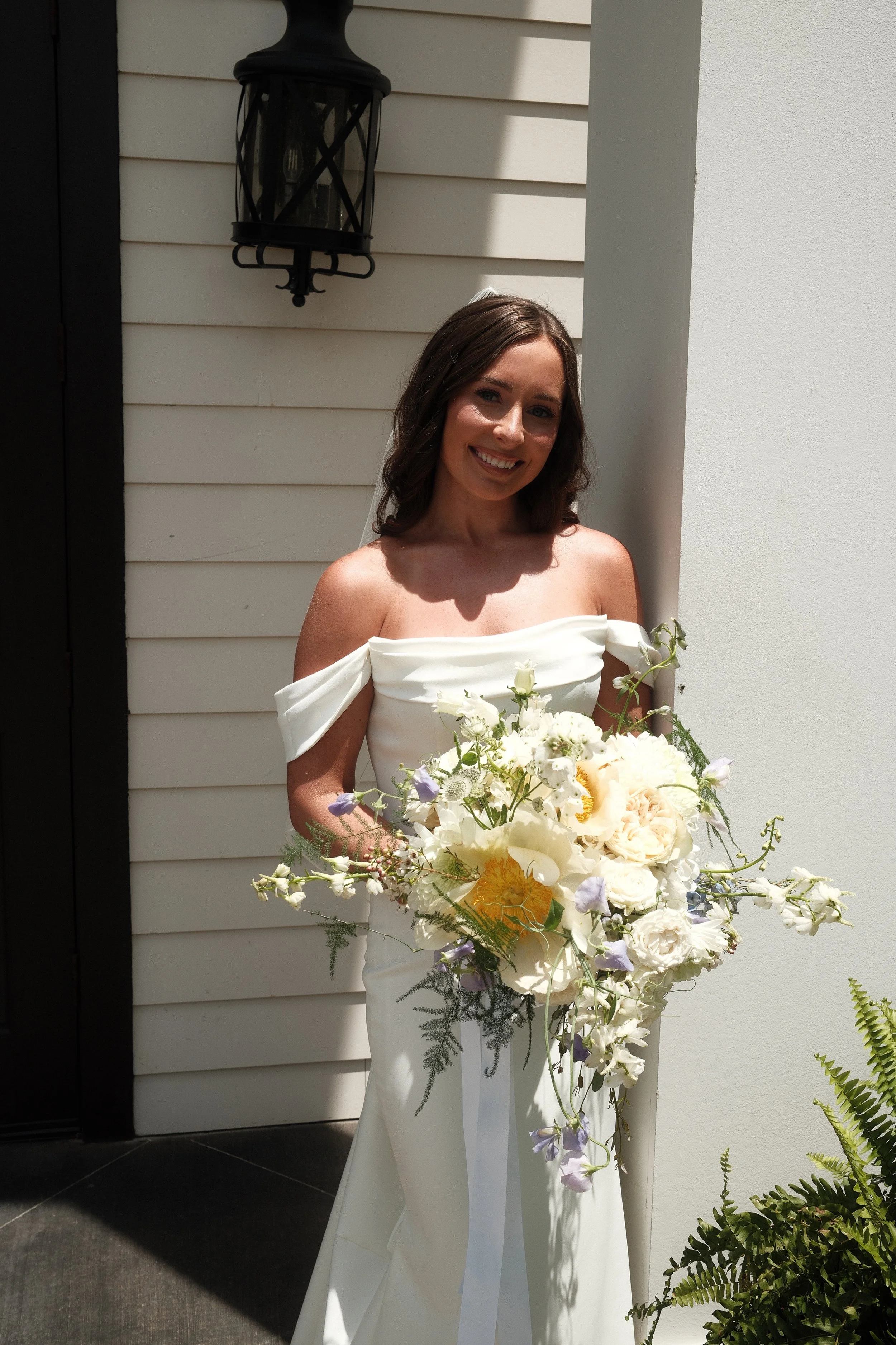 A smiling woman dressed in a white off-shoulder gown holding a large bouquet of white and pale purple flowers in front of a white wall with a black lantern-style outdoor light fixture.