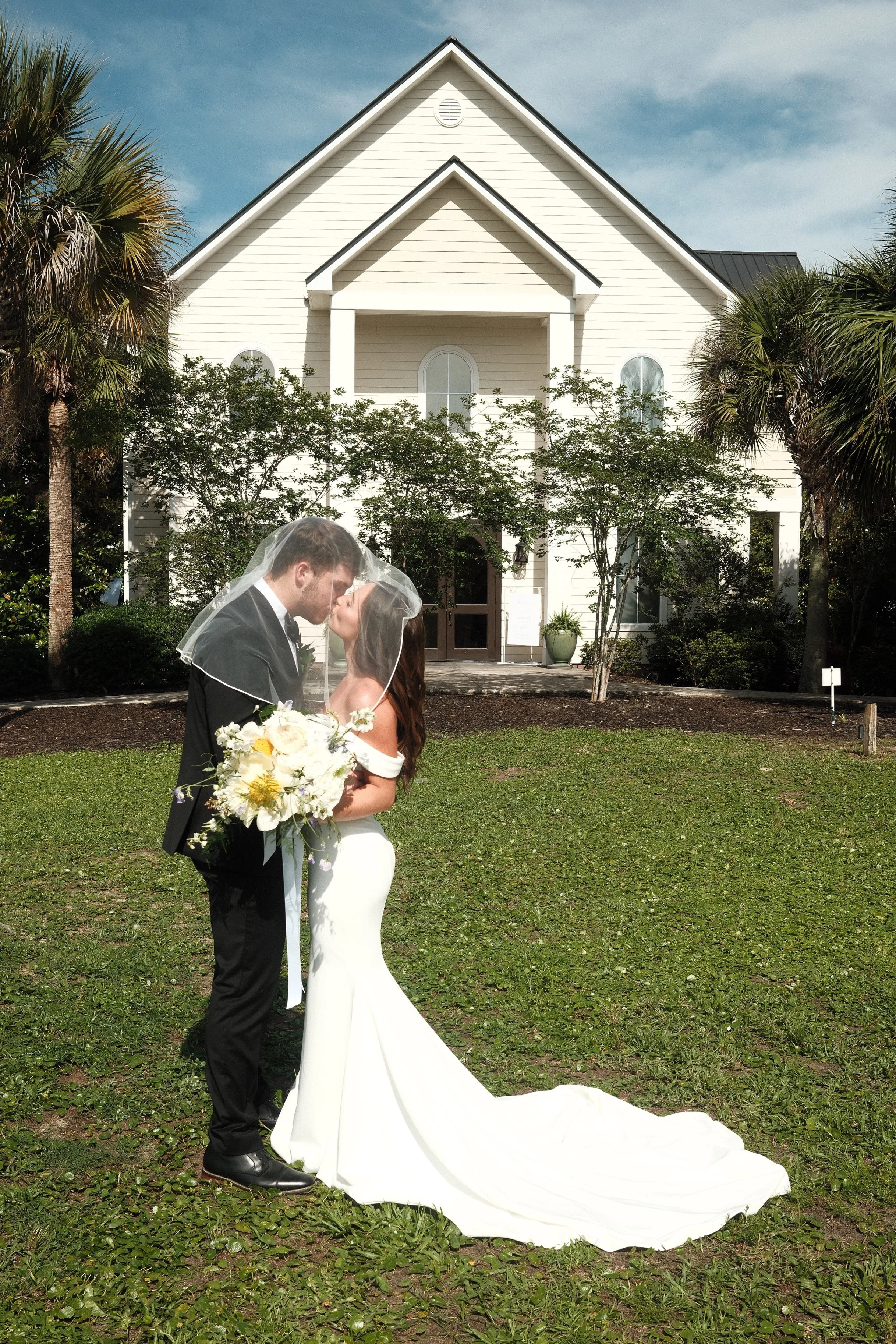 A bride and groom share a kiss under a wedding veil outside a white house with a steep roof, surrounded by green trees and grass.