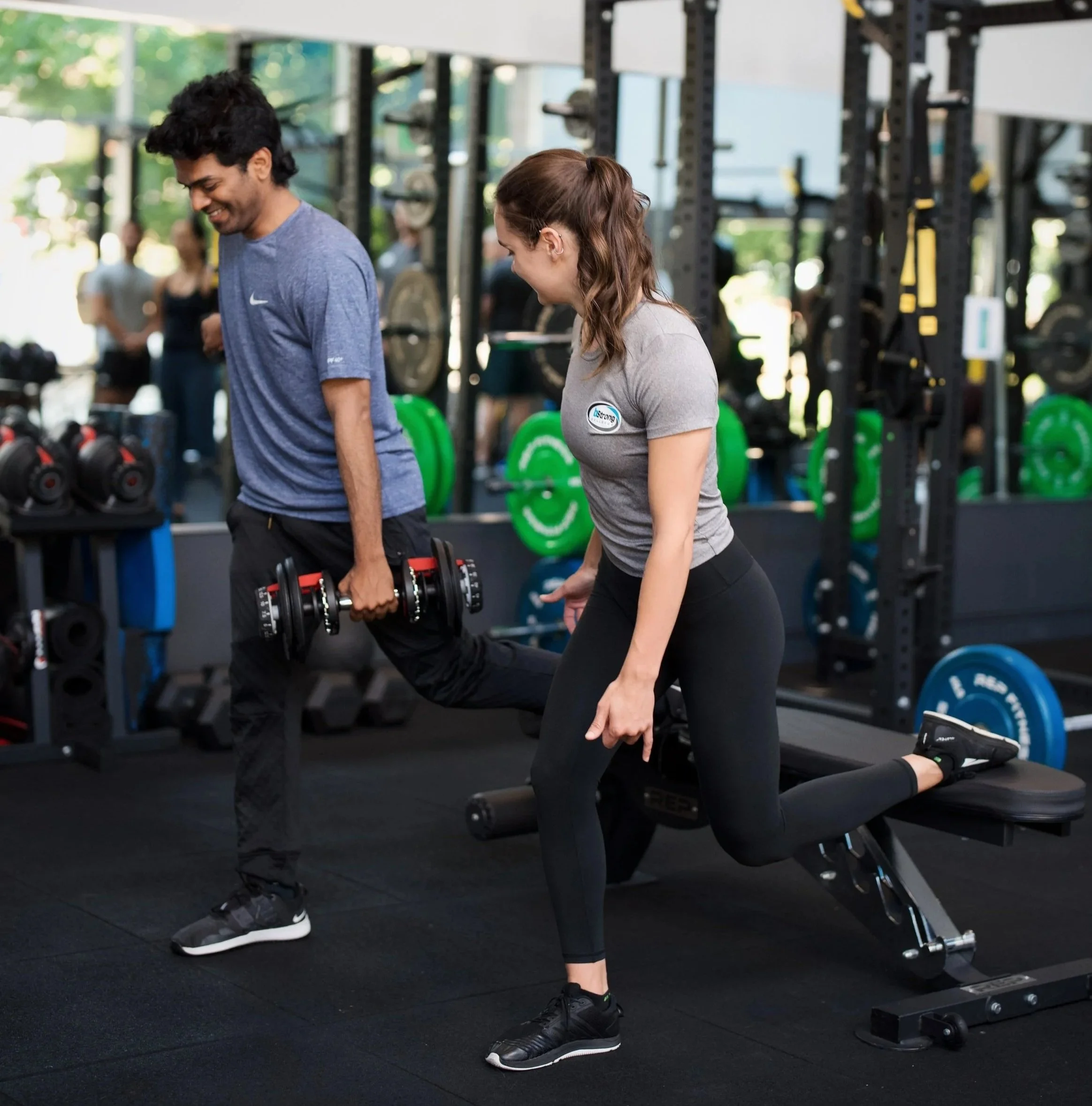 A man and a woman exercising together in a gym, both lifting dumbbells while standing. The gym has weightlifting equipment, green weight plates, and other members in the background.