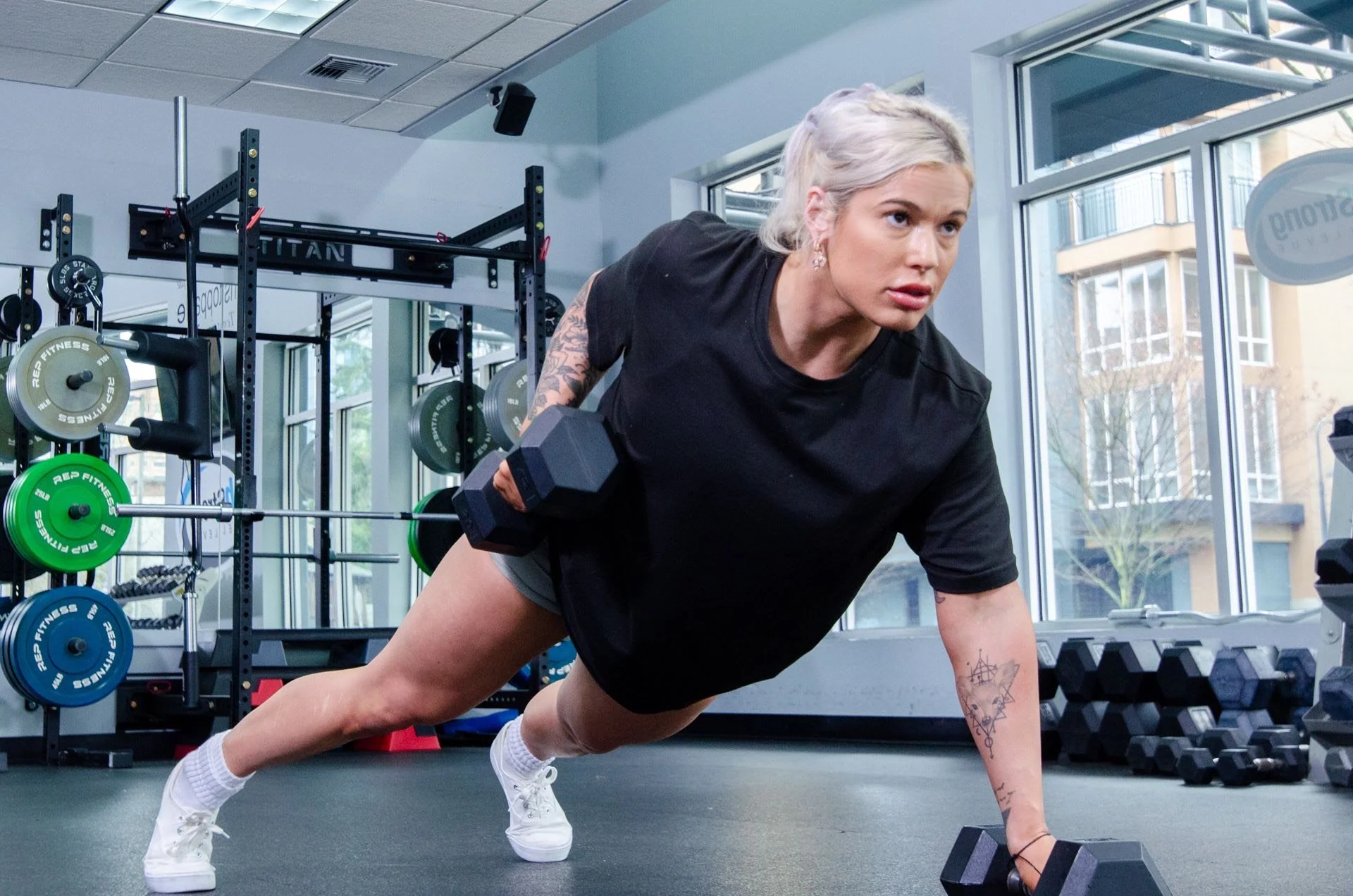 A woman with tattoos doing a plank exercise in a gym, holding a dumbbell in one hand, with gym equipment and weights around her.