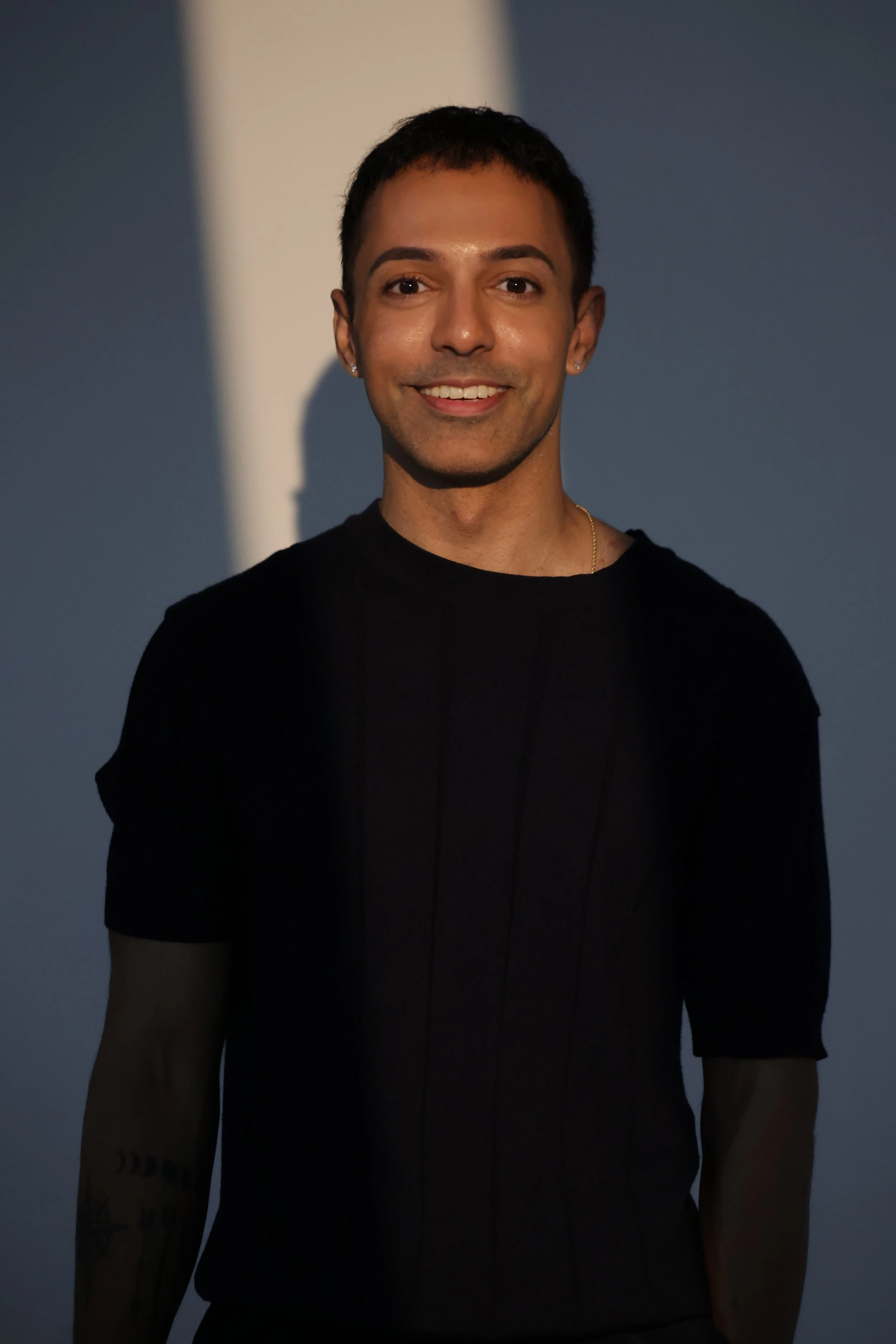 Front view of a young man with short dark hair, wearing a black shirt, earrings, and a gold necklace, smiling against a background with soft lighting and shadows.