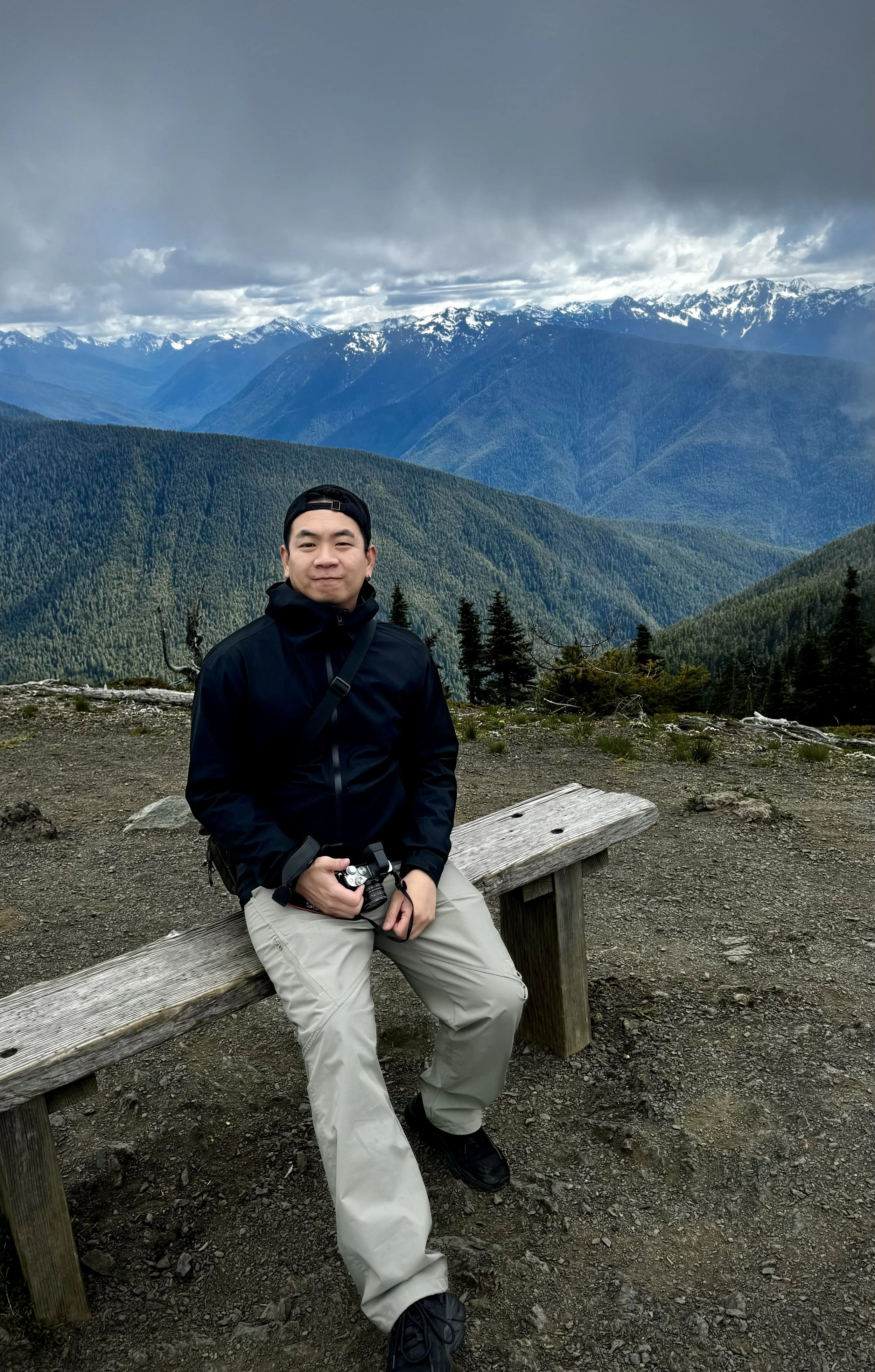 A man sitting on a wooden bench in a mountainous area with green forested mountains and snow-capped peaks in the background, under a cloudy sky.