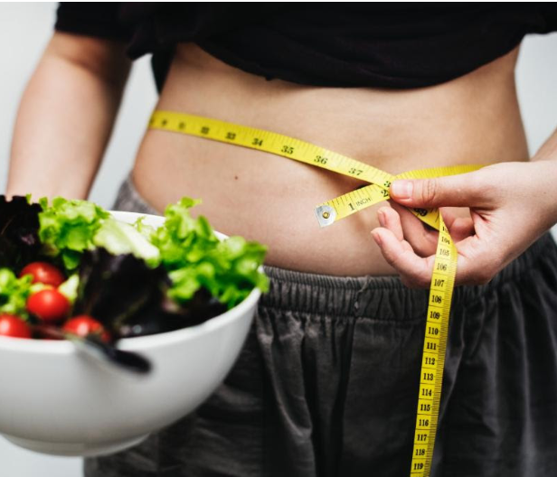 A person holding a salad in one hand and trying to hold a tape measure to measure their waist circumference in the other
