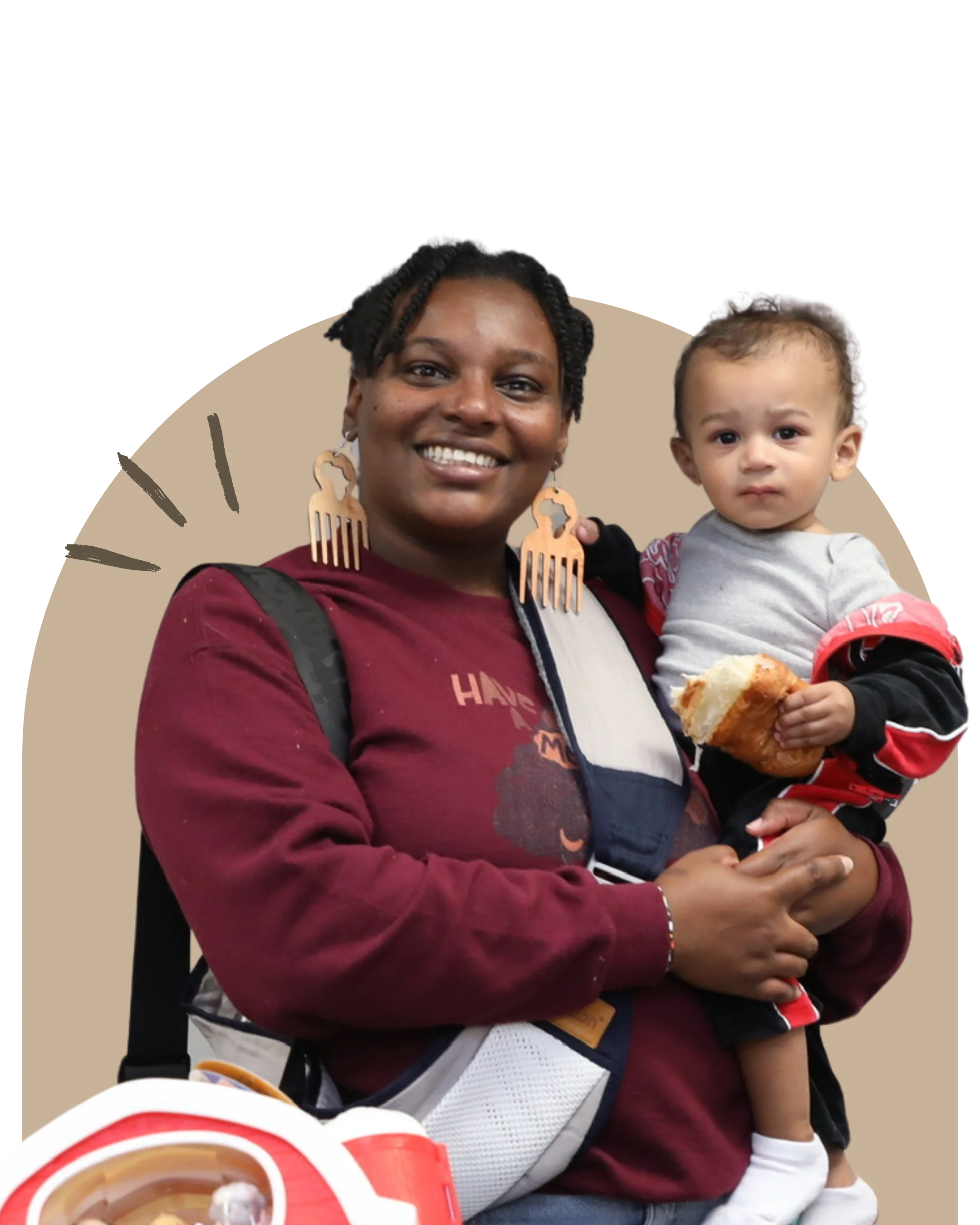 A smiling woman holding a young child while standing in front of a beige background. The woman wears earrings and a maroon hoodie, and the child wears a light gray shirt with red and black accents and holds a piece of bread. There are food items at the bottom of the image.