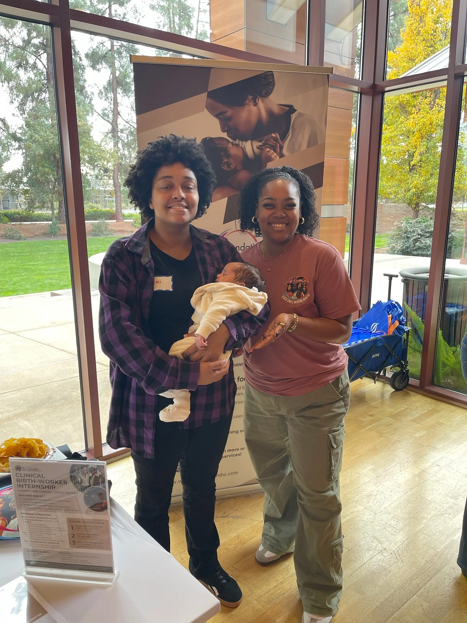 Two women, one holding a newborn baby, stand inside a building near large windows with trees outside. Behind them is a poster of a mother and child, and a table with informational flyers about a clinical birth-worker internship.