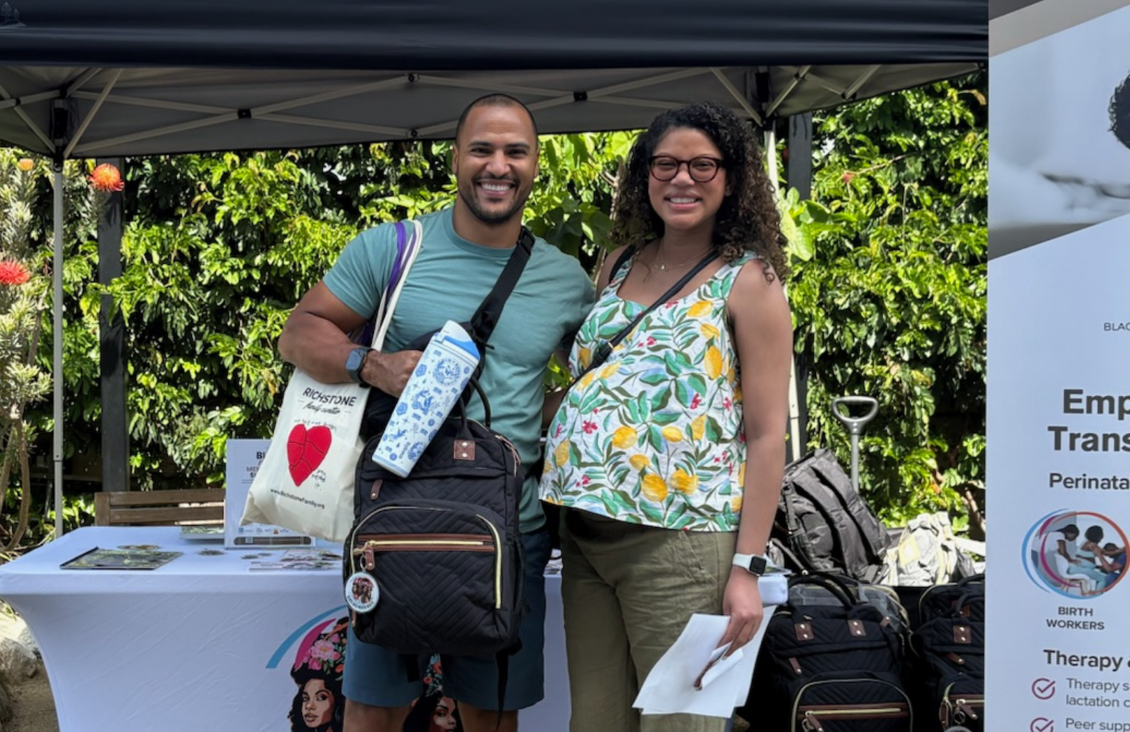 Two smiling people, a man and a woman, standing outdoors in front of a table and greenery, at an event booth with informational posters. They are holding bags and wearing casual clothes.