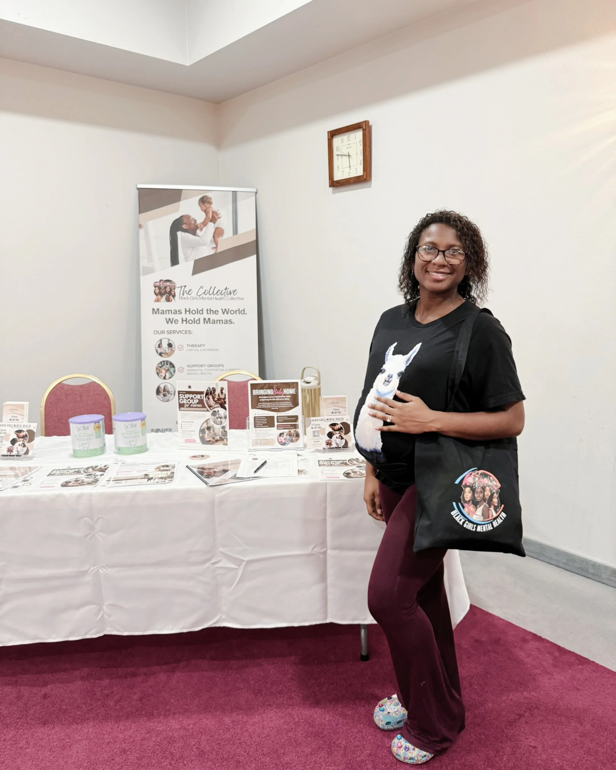 A woman smiling and standing in front of a table displaying brochures and information materials at an indoor event. She is wearing glasses, a black t-shirt with a graphic, maroon pants, and colorful fuzzy slippers. Behind her is a banner for The Collective and a wall clock.