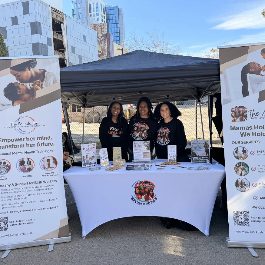 Three women standing behind a table at an outdoor event under a black canopy, with two informational banners on either side promoting mental health services for Black girls, with a cityscape in the background.