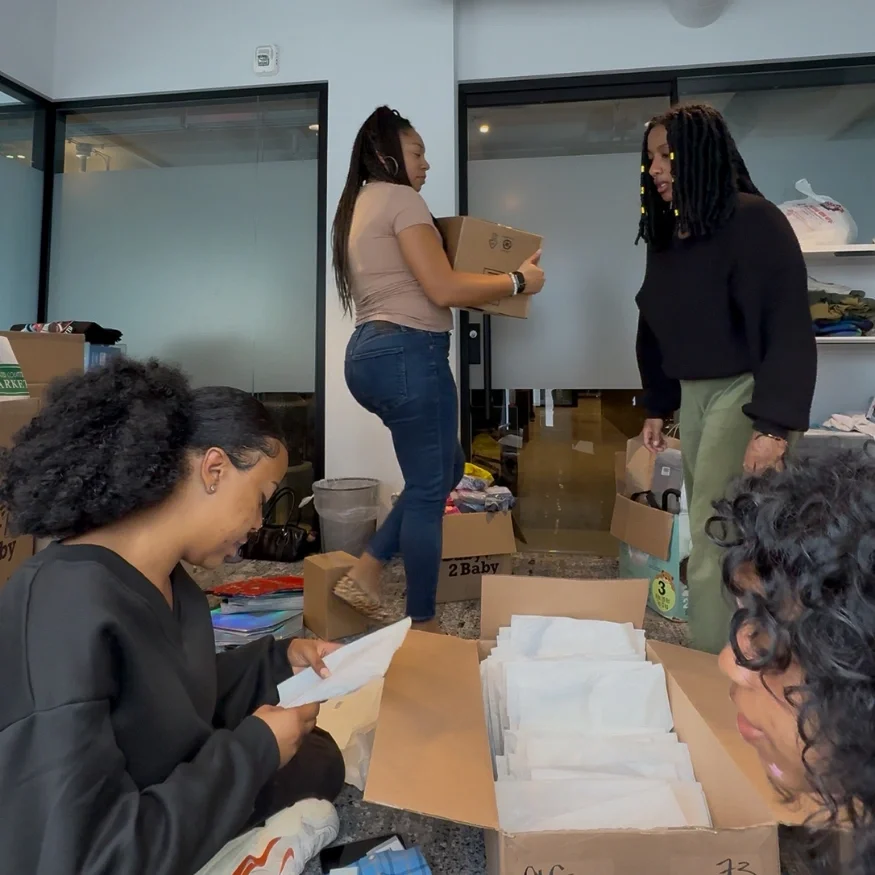 Four women are organizing and packing documents or items in a room filled with boxes and supplies.