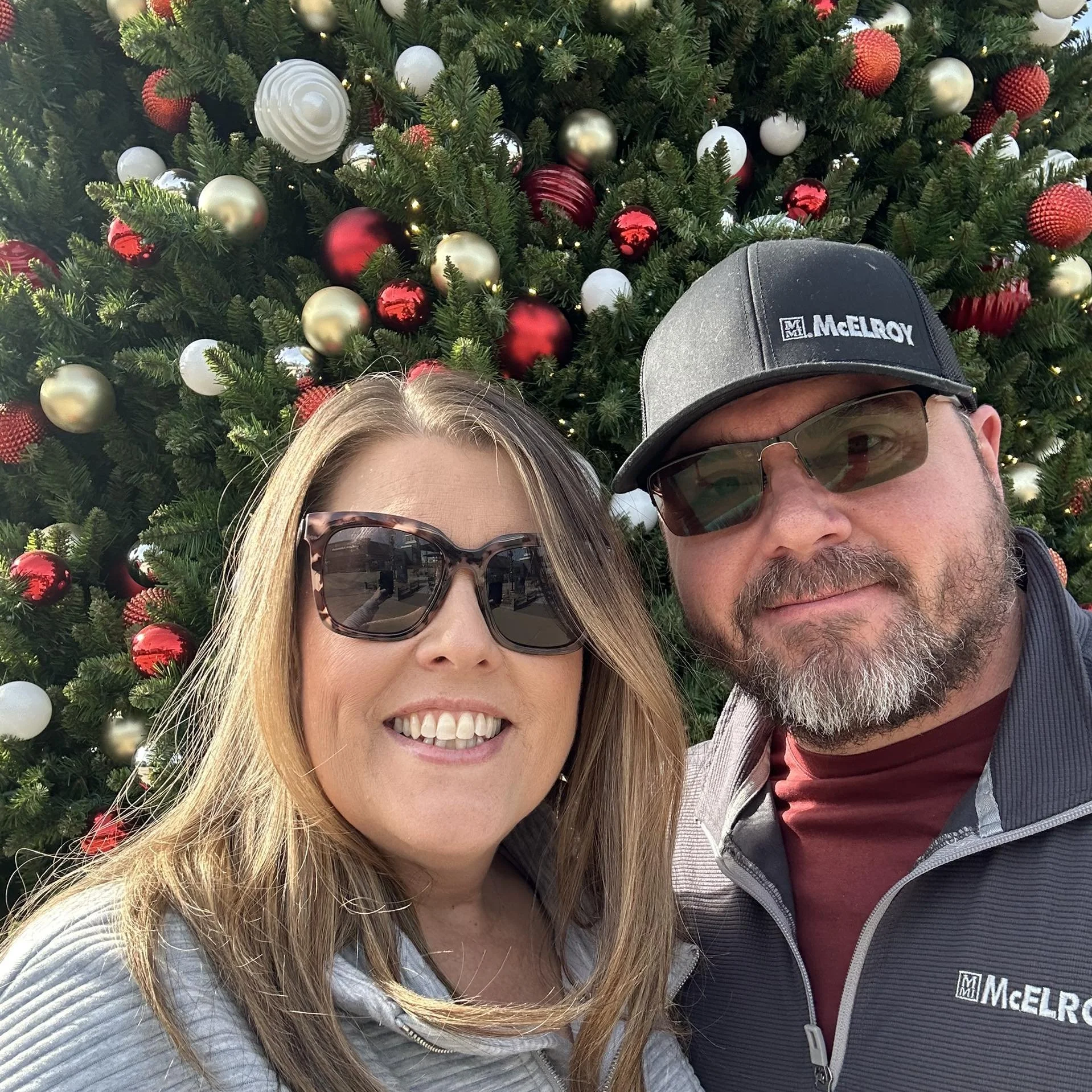 A smiling woman wearing sunglasses and a man wearing sunglasses and a baseball cap, both standing in front of a decorated Christmas tree with red, gold, and white ornaments.