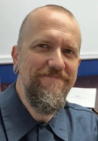 A man with a beard and mustache smiling, wearing a dark collared shirt, in an indoor setting with a light-colored wall and a blue object in the background.