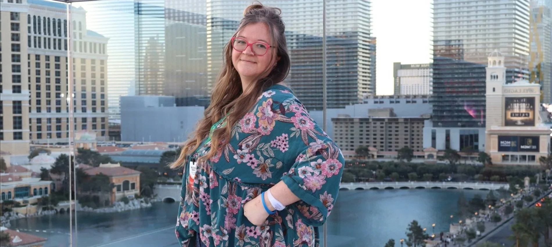 Young woman with long wavy hair, glasses, and a floral shirt, standing with hands in pockets, smiling outdoors with a cityscape and waterway in the background.