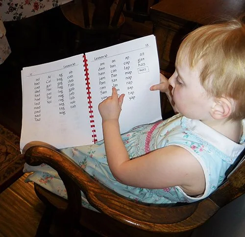 A young child learning to read with Alpha Phonics. She is seated in a chair and pointing at the word she is reading.