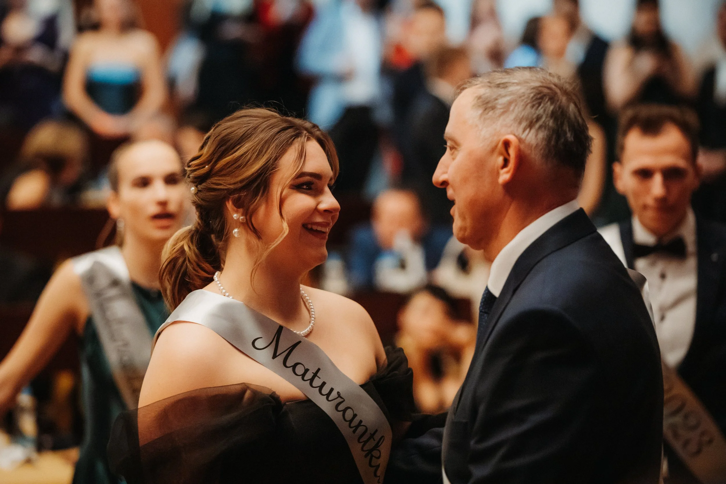 Young woman at a formal event wearing a sash with "Maturantky" text, smiling and interacting with a man in a suit.