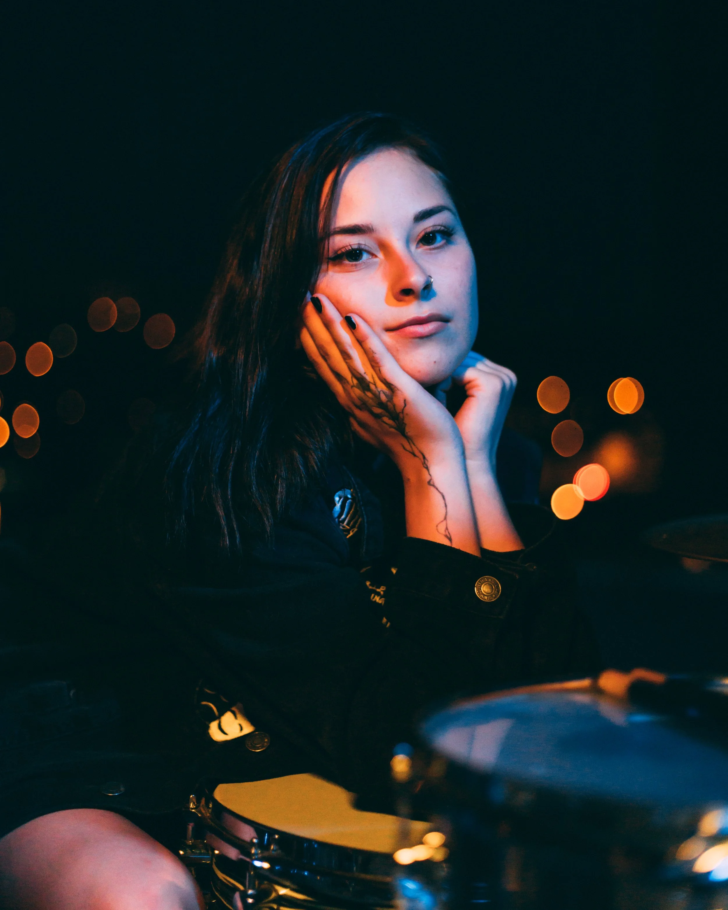 Person with dark hair, resting their chin on hands, with facial piercings and tattoos, in dim lighting.