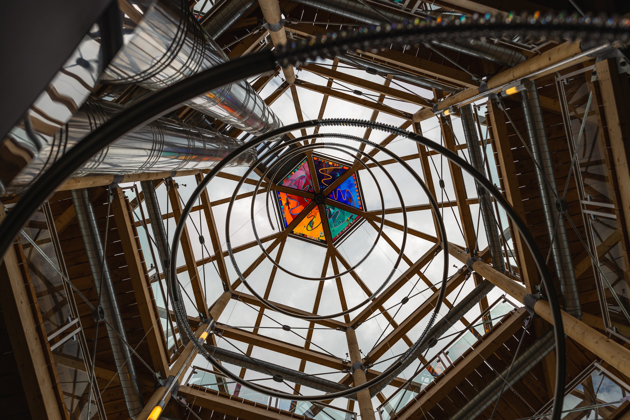 View looking up at a geometric wooden and metal architectural structure with a colorful stained glass panel at the top center.