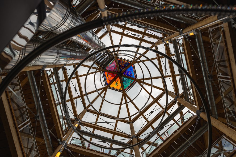 View looking up at a geometric wooden and metal architectural structure with a colorful stained glass panel at the top center.