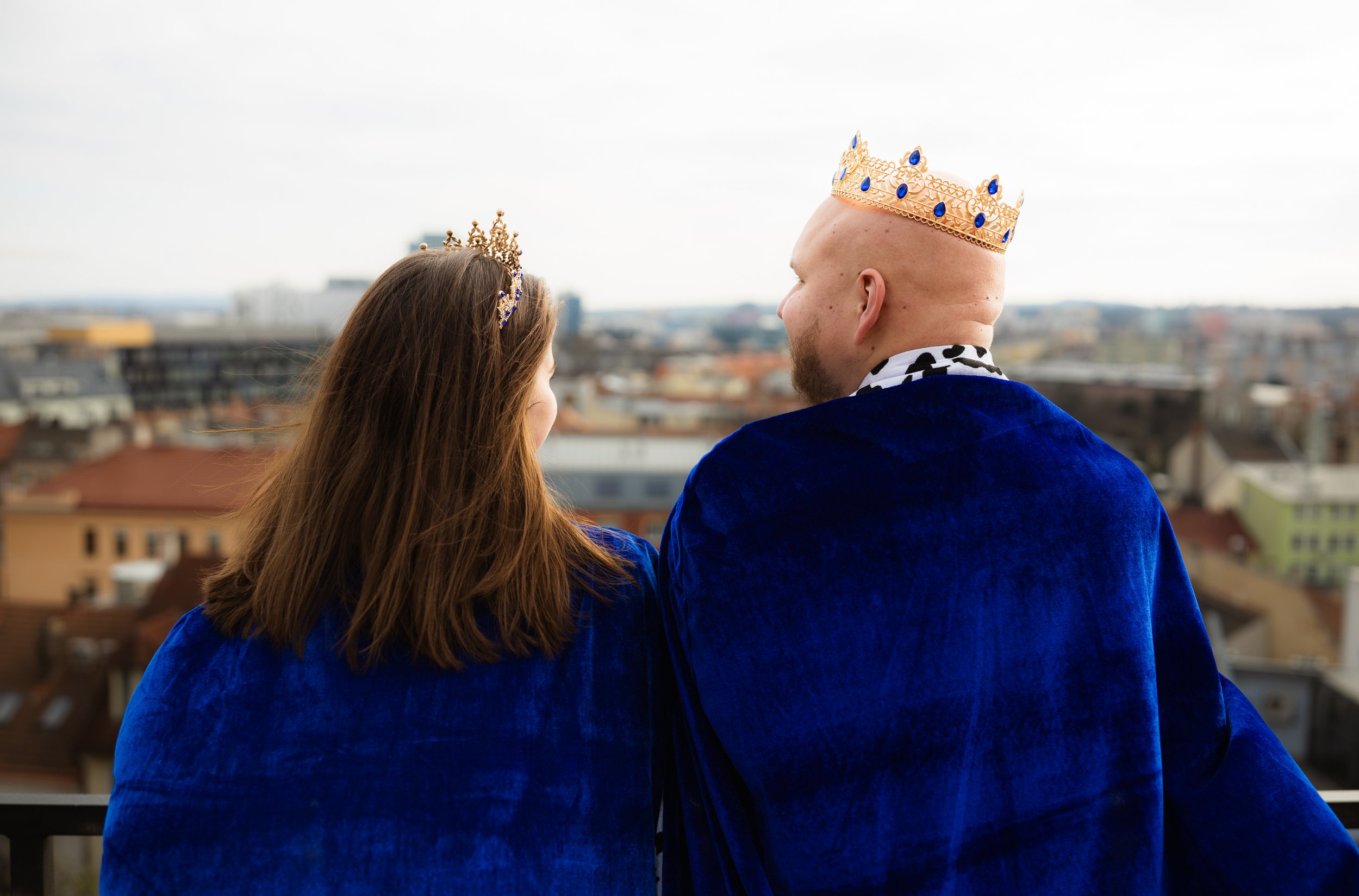 Two people wearing crowns and royal blue cloaks, looking over a cityscape from a balcony.