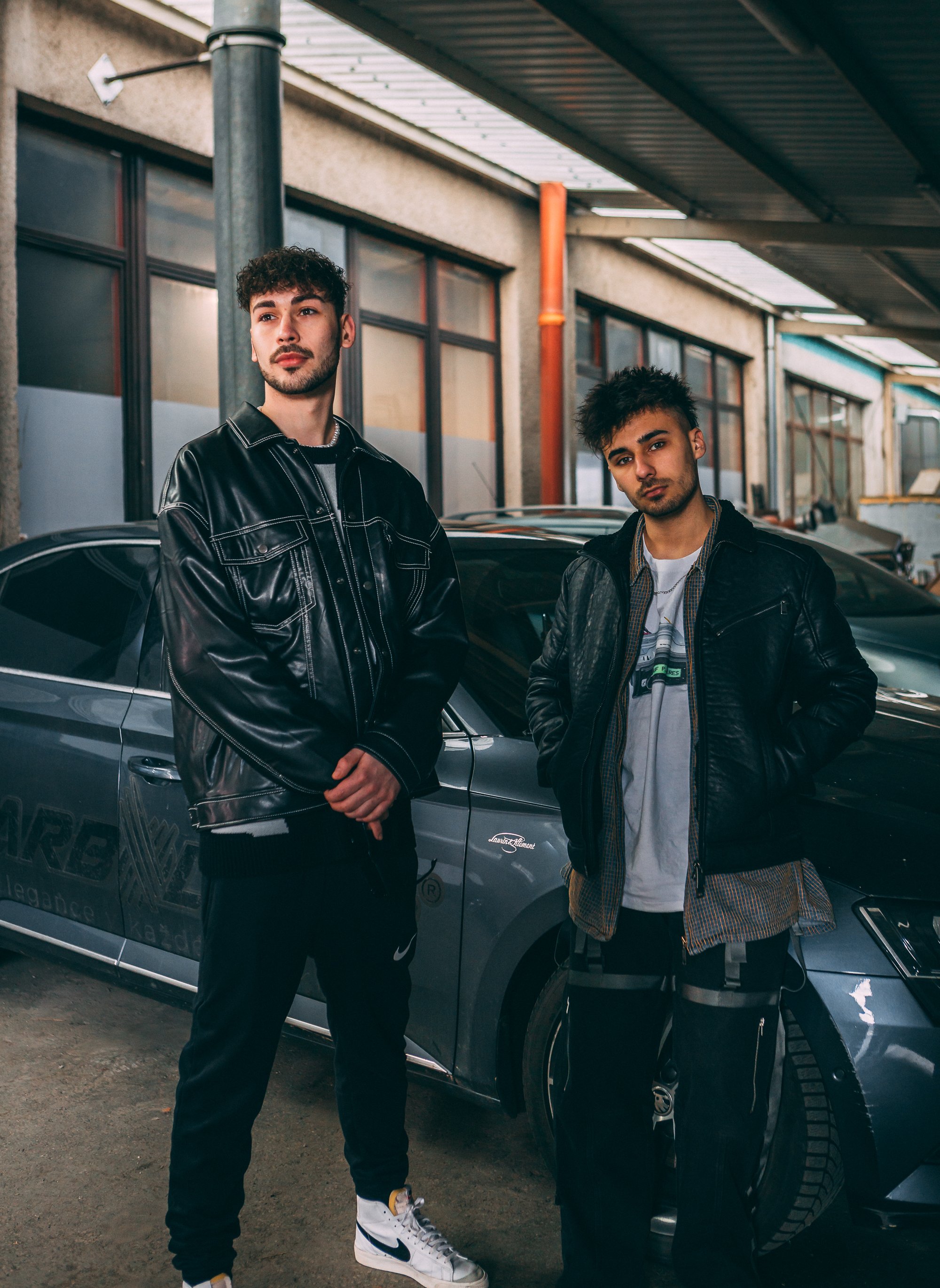 Two young men in casual urban outfits standing next to a car in a garage or industrial setting.