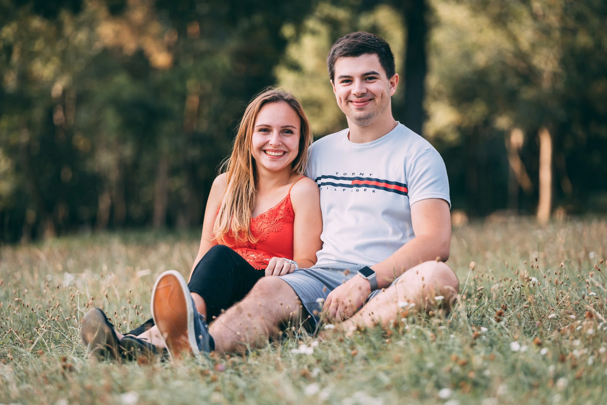 A smiling couple sitting on grass in a park, with trees in the background; the woman wears a red top, and the man wears a light-colored T-shirt and shorts.