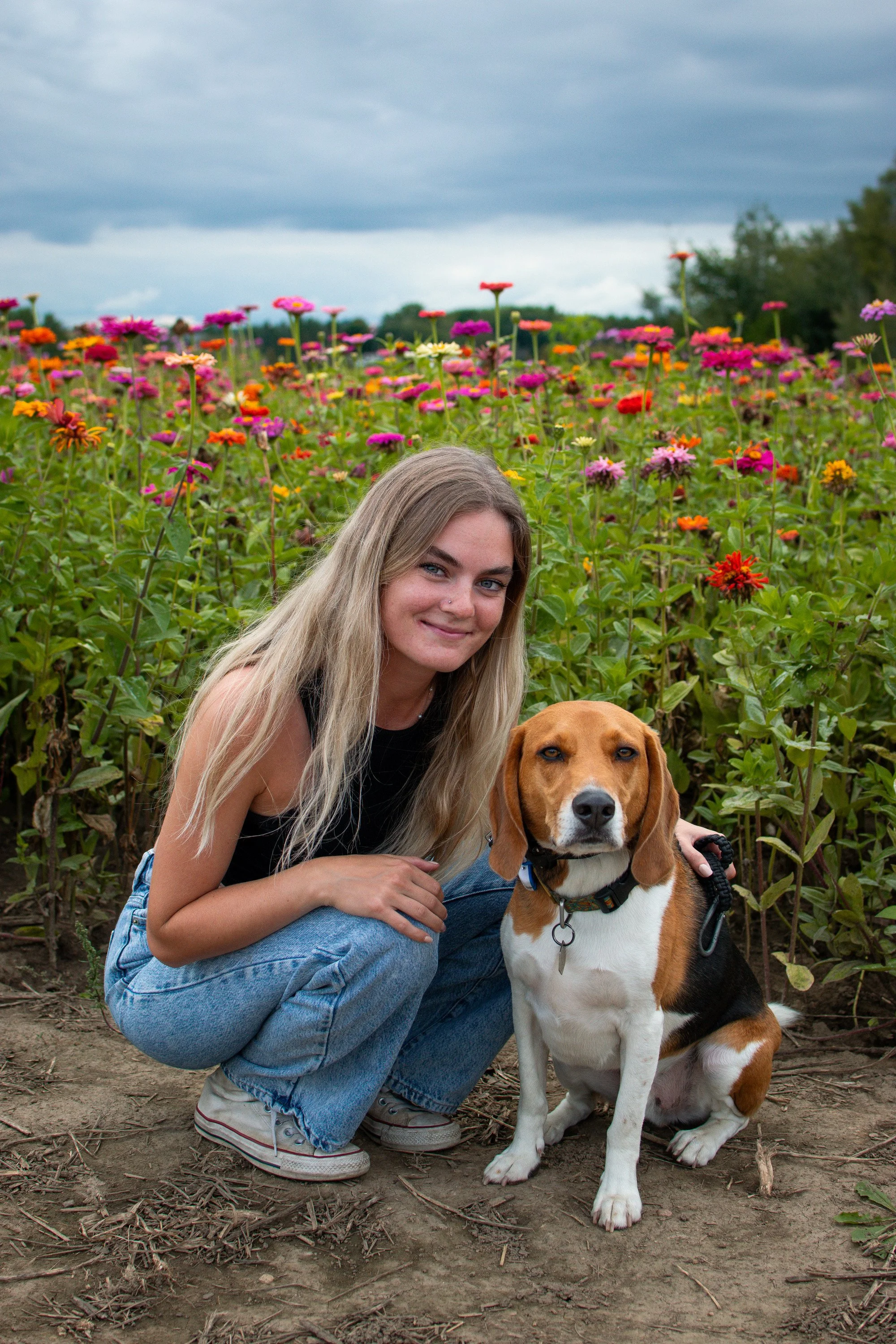A young woman with long blonde hair crouches next to a beagle dog in a colorful flower field, under a cloudy sky.