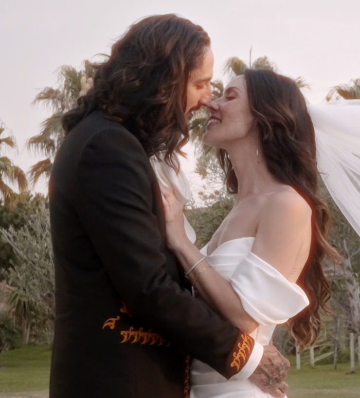 Un couple en tenue de mariage s'embrasse tendrement en extérieur, entouré de végétation et de palmiers, lors d'un coucher de soleil.