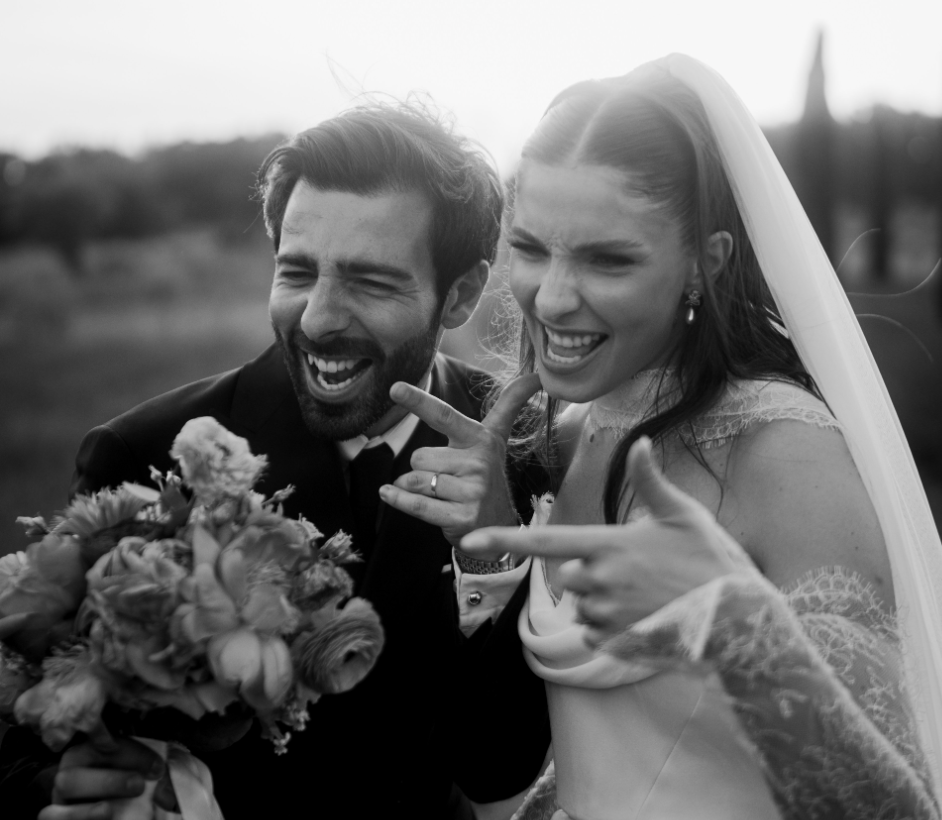 Un couple de mariés fait une photo amusante, la femme porte une robe de mariage avec voilette et le marié un costume, ils sourient et pointent leurs doigts vers l'objectif, avec un bouquet de fleurs dans la main.