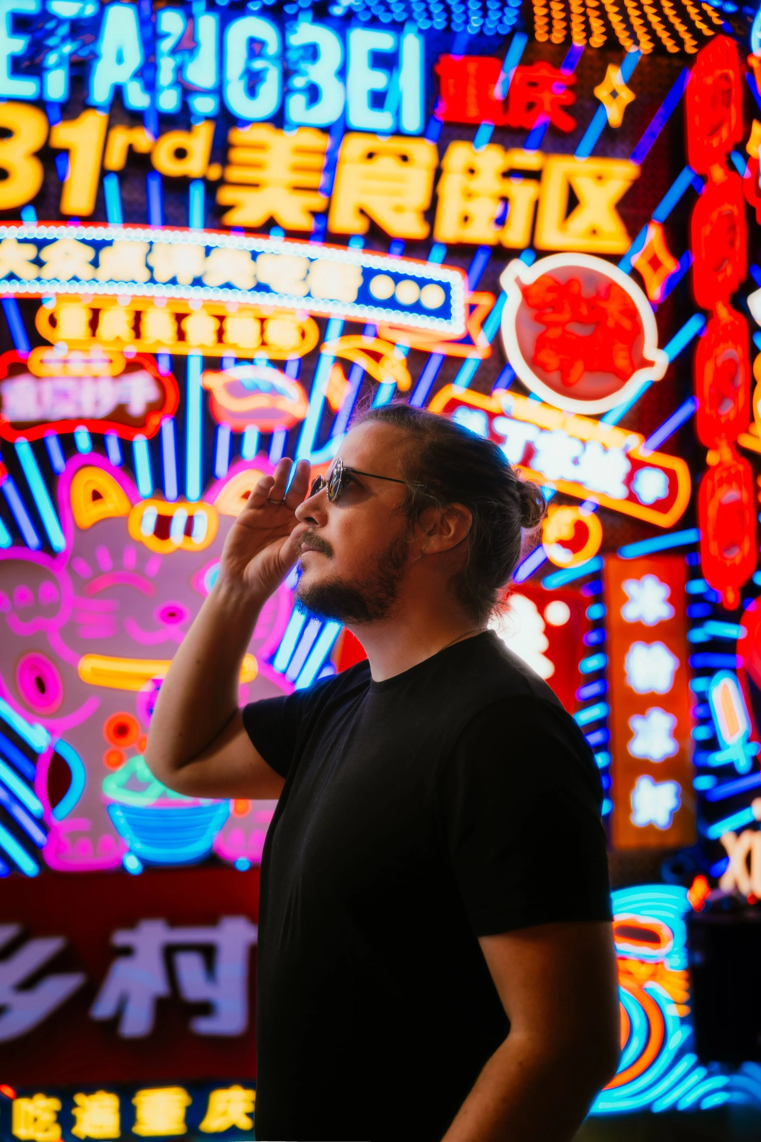 Homme avec lunettes de soleil, ayant une barbe et des cheveux longs attachés, pose devant un fond de panneaux lumineux de style néon en caractères chinois et japonais avec des motifs colorés, dans une ambiance urbaine nocturne.
