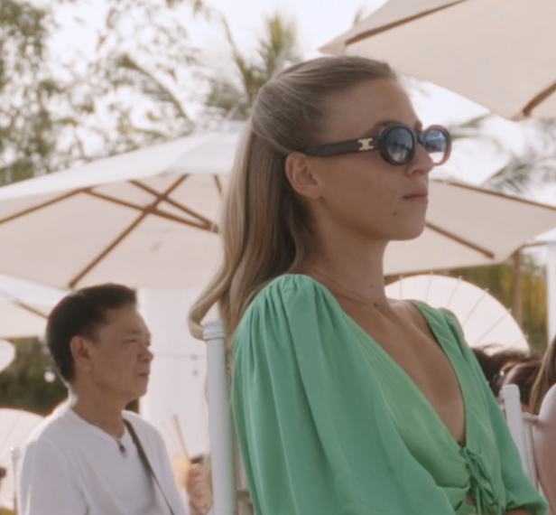 Une femme portant des lunettes de soleil et une robe verte assise sous un parasol, avec une autre personne en arrière-plan.