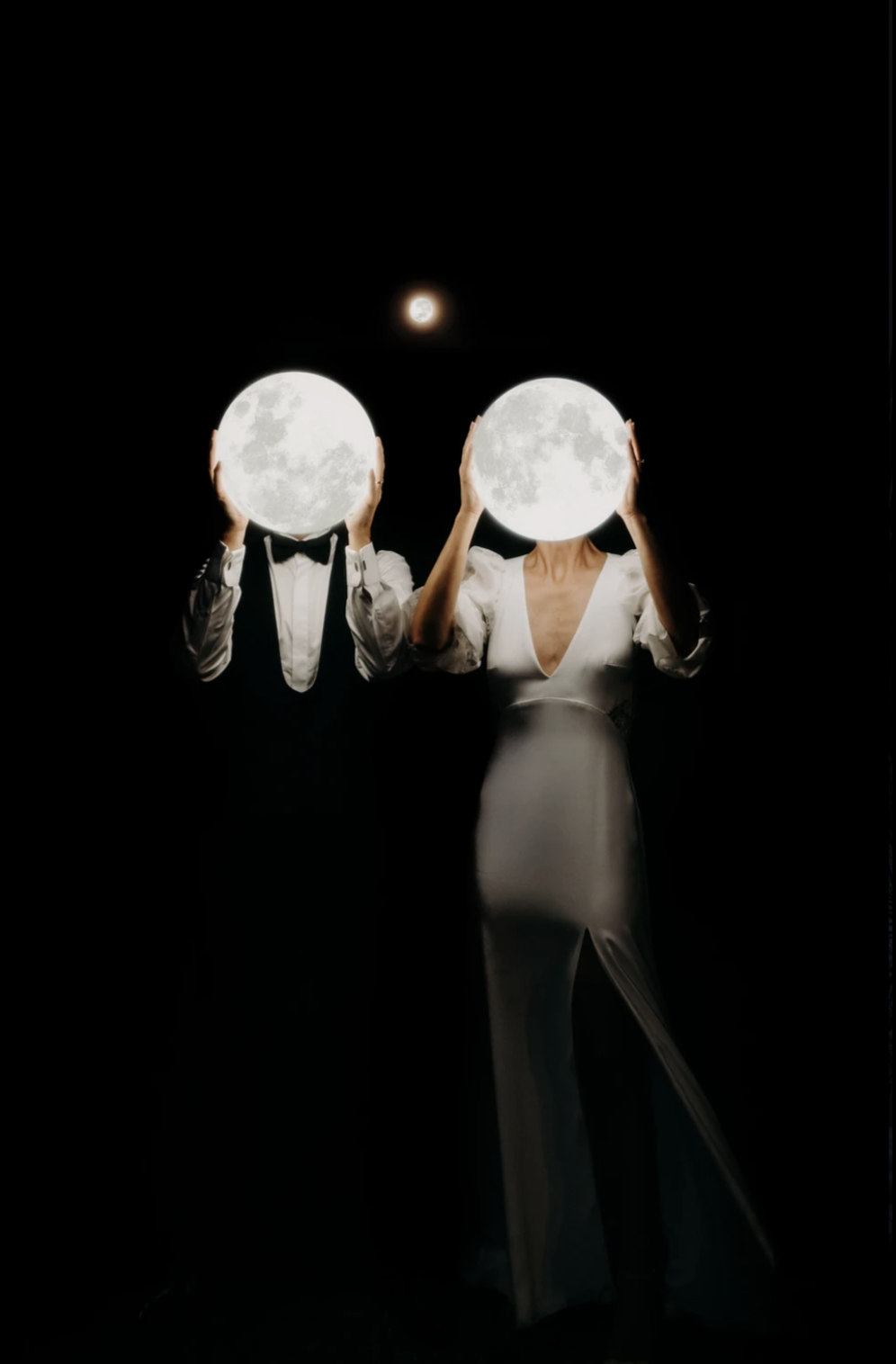 Deux personnes habillées élégamment, l'une avec un costume noir avec un nœud papillon, l'autre en robe blanche, tiennent des lampes en forme de lune devant leur visage, créant une illusion de lune dans un ciel noir, avec la pleine lune en arrière-plan.