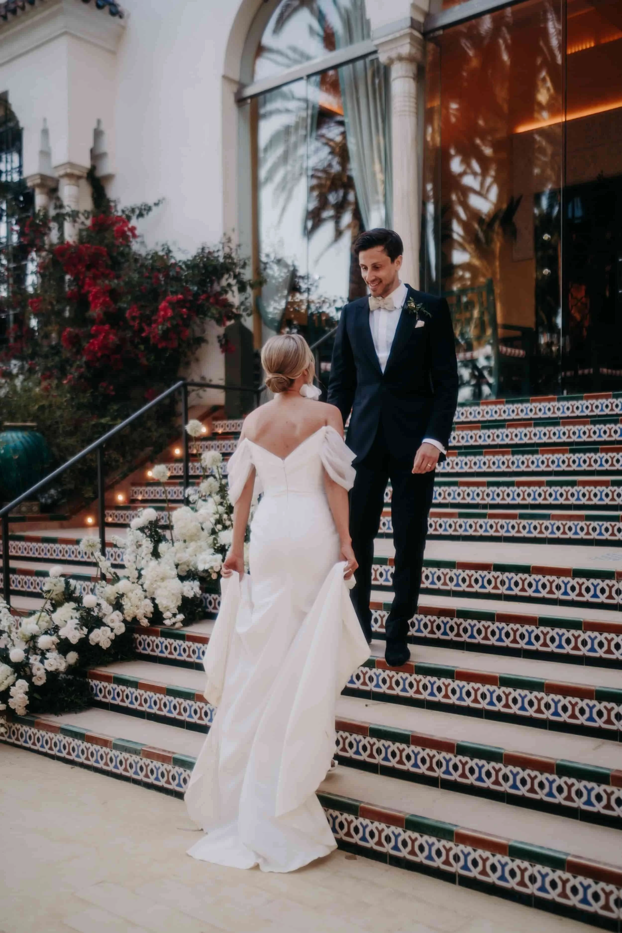 Un couple de mariés descend un escalier décoré de fleurs blanches devant un bâtiment avec de grandes fenêtres et des plantes tropicales en arrière-plan.