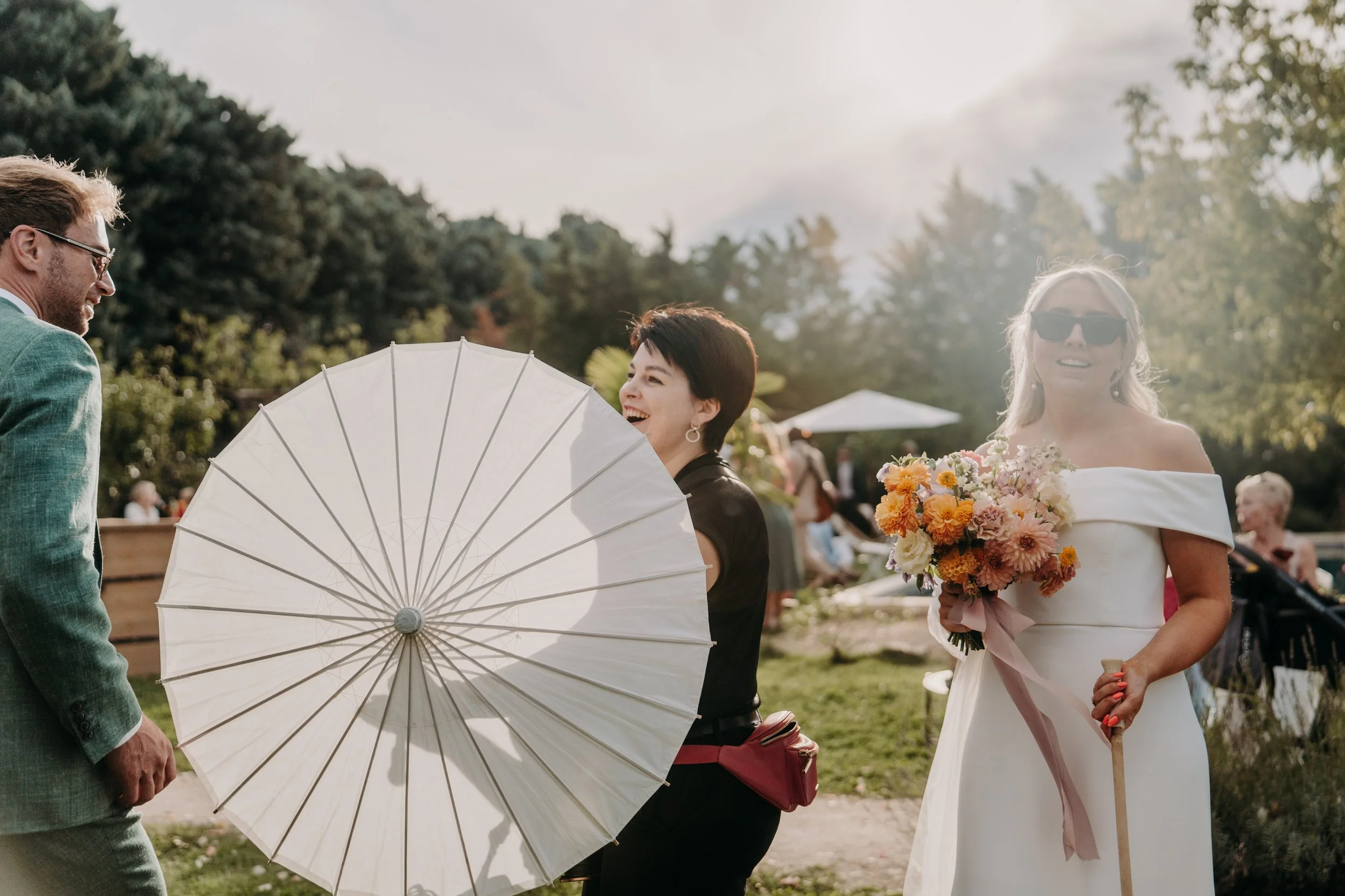 Trois personnes lors d'une célébration en plein air, avec un homme, une femme avec un parapluie et une autre femme en robe blanche et lunettes de soleil, tenant un bouquet de fleurs.