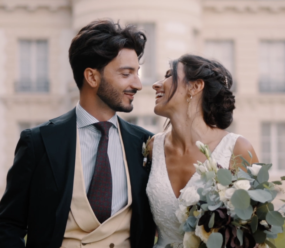 Un couple souriant, probablement lors d'un mariage, avec la femme tenant un bouquet de fleurs et portant une robe blanche, et l'homme en costume avec une cravate, devant un bâtiment ancien.