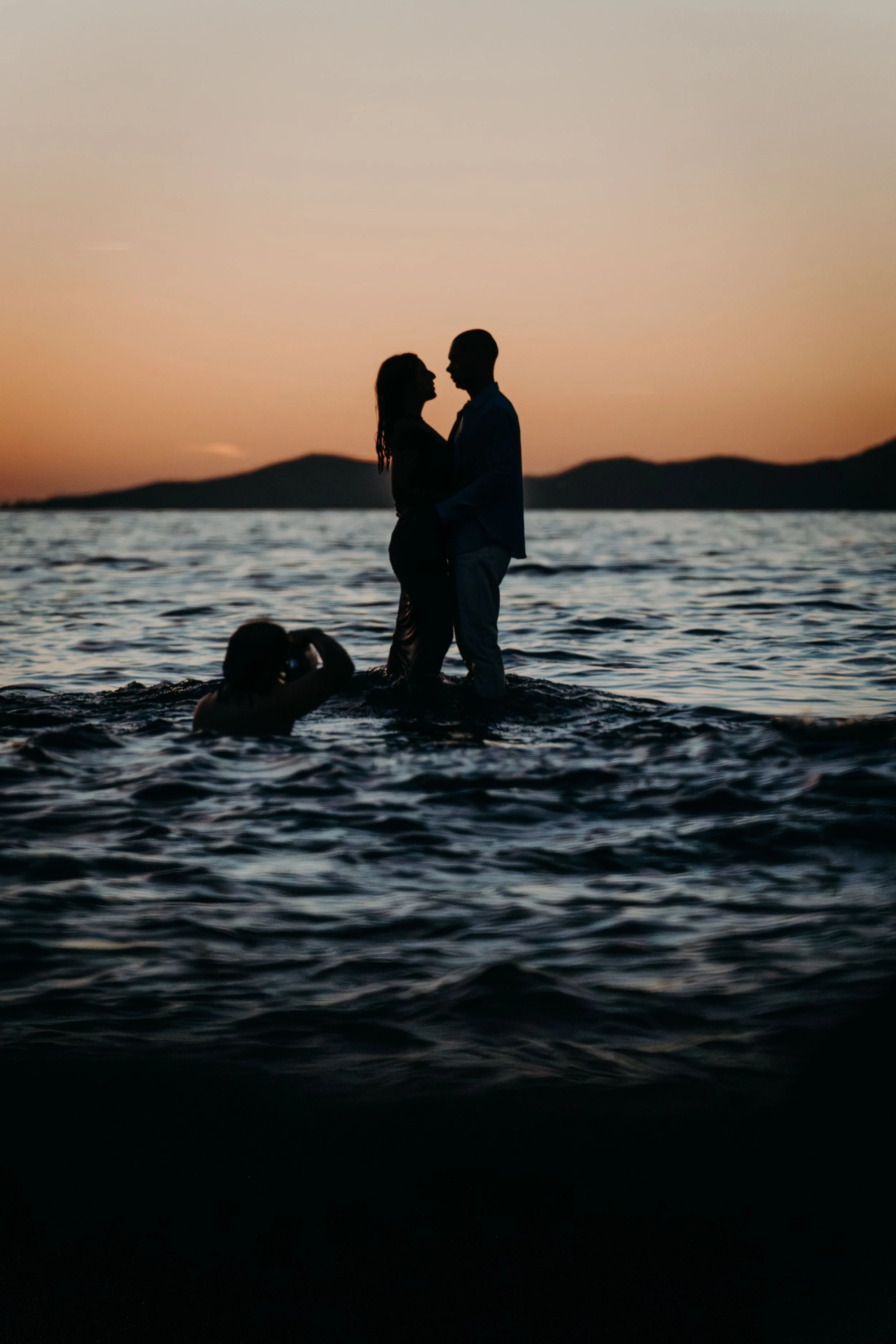 Un couple se tenant par la main dans l'eau au coucher du soleil, avec une personne sous l'eau prenant une photo.