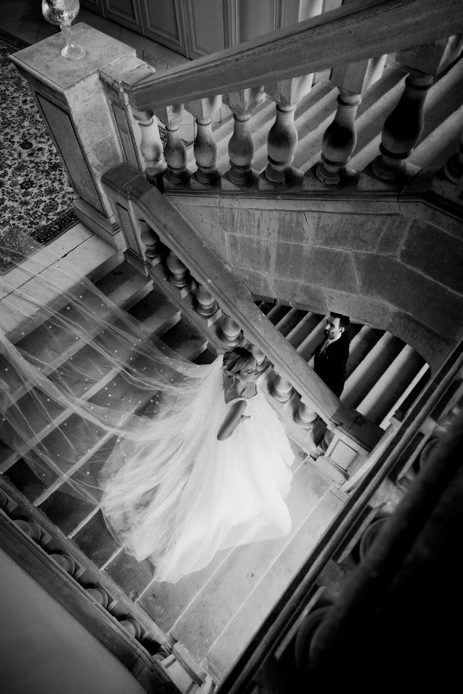 couple-maries-escalier-chateau-voile-envol-noir-blanc-photographe-mariage-film-mariage-the-wild-strawberry.webp